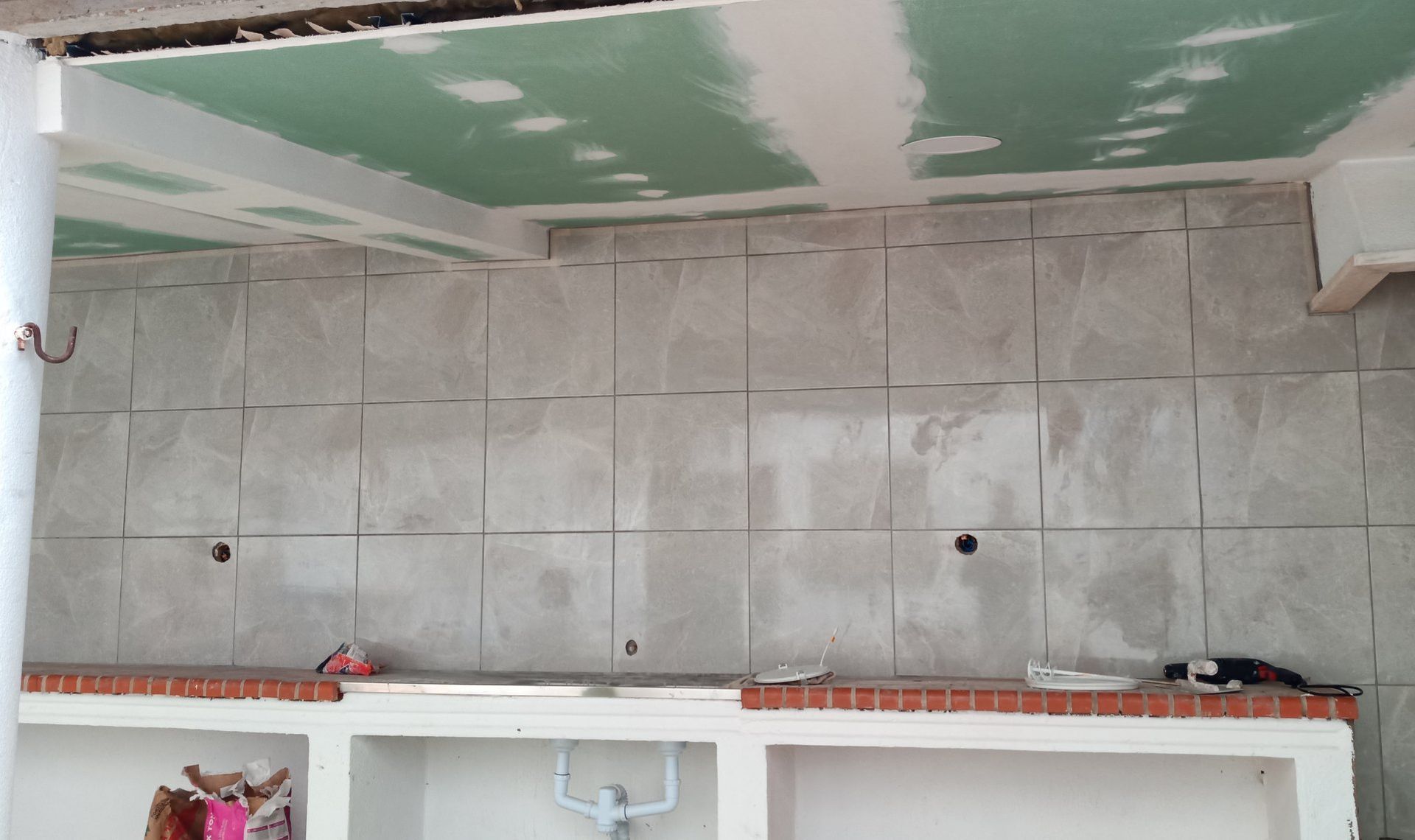 Quarto em reforma, com teto de gesso acartonado e parede revestida com azulejos. É possível ver uma bancada com tijolos e prateleiras brancas.