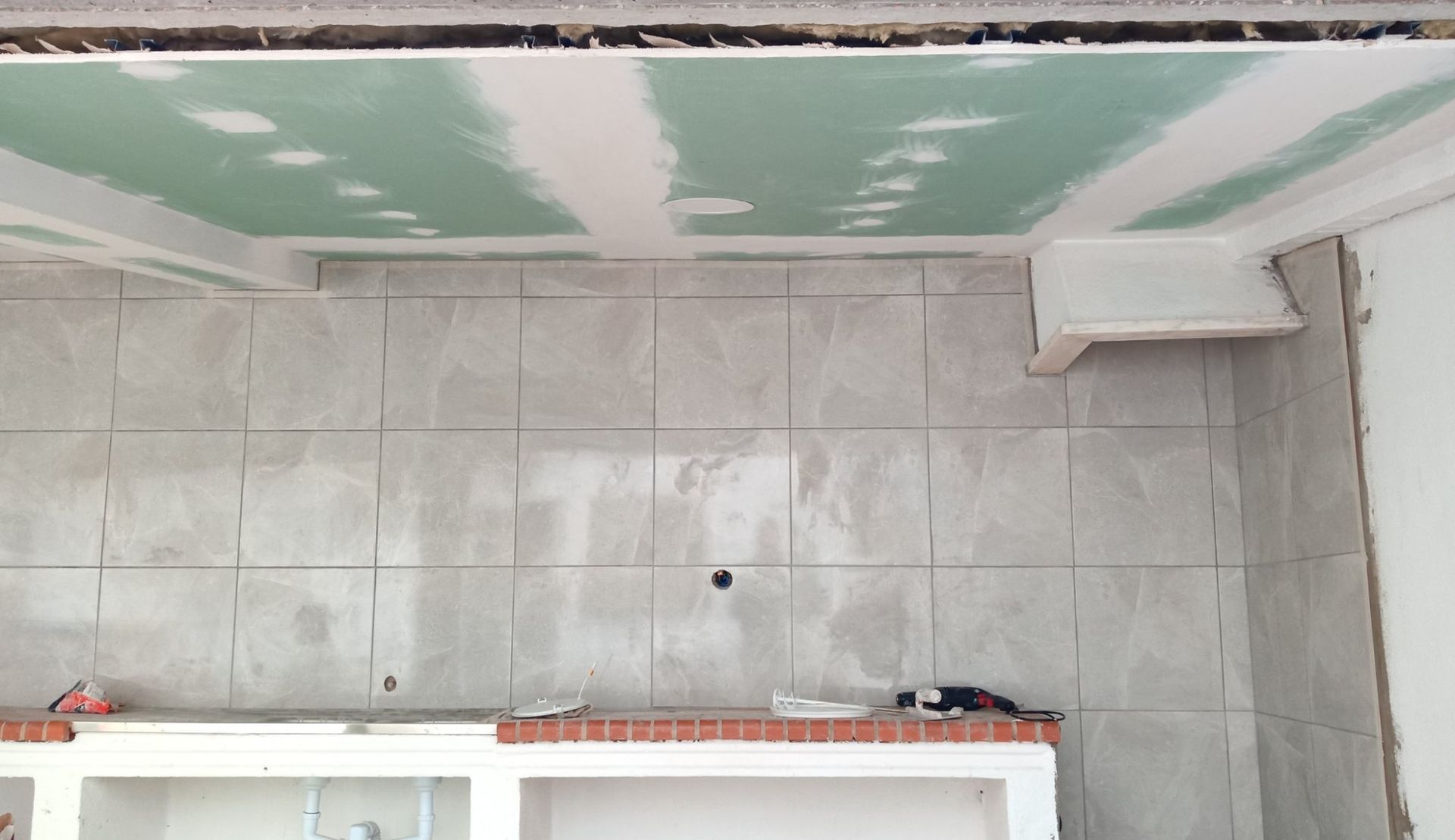 Construção em andamento: parede revestida com azulejos, teto inacabado com drywall e gesso. Plataforma de tijolos abaixo.