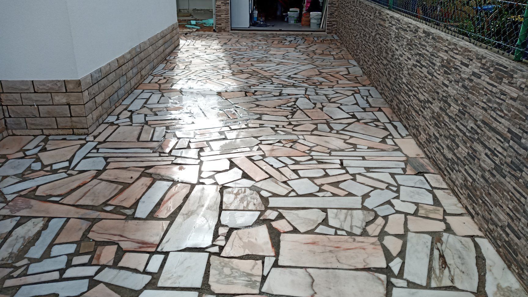 Entrada de veículos pavimentada com pedras claras de formato irregular, delimitada por muros de tijolo e pedra texturizada.