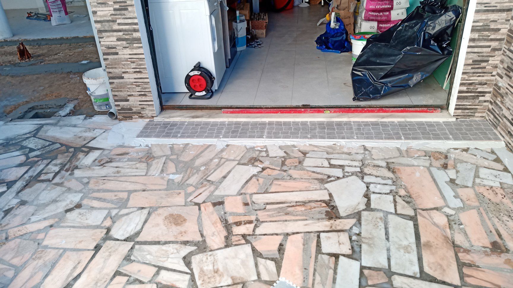 Entrada da loja com piso de ladrilho. Uma porta dá acesso ao interior, onde vários itens estão visíveis. Um cabo de extensão vermelho também está à vista.
