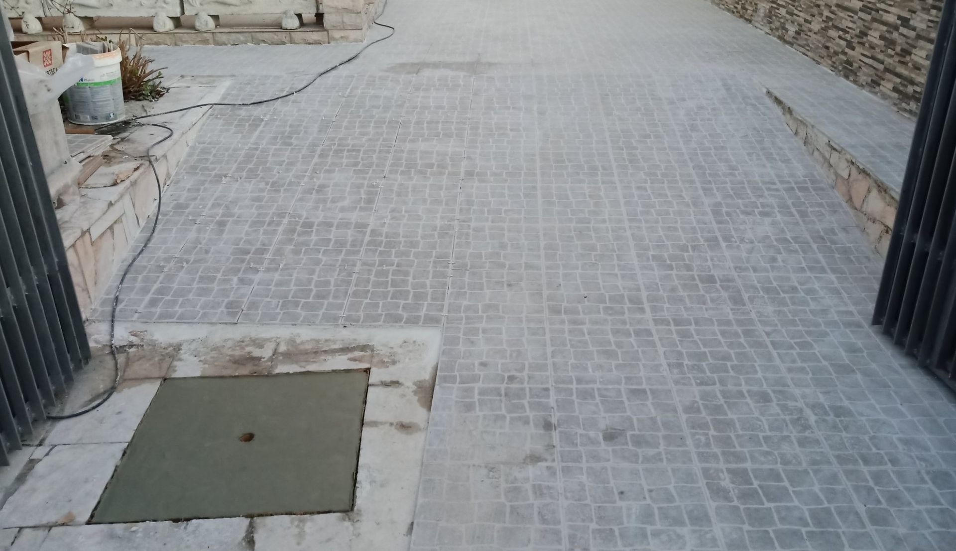 Área de pátio pavimentada com tijolos cinza, com uma tampa metálica quadrada no canto inferior esquerdo e um portão metálico parcialmente aberto à direita.