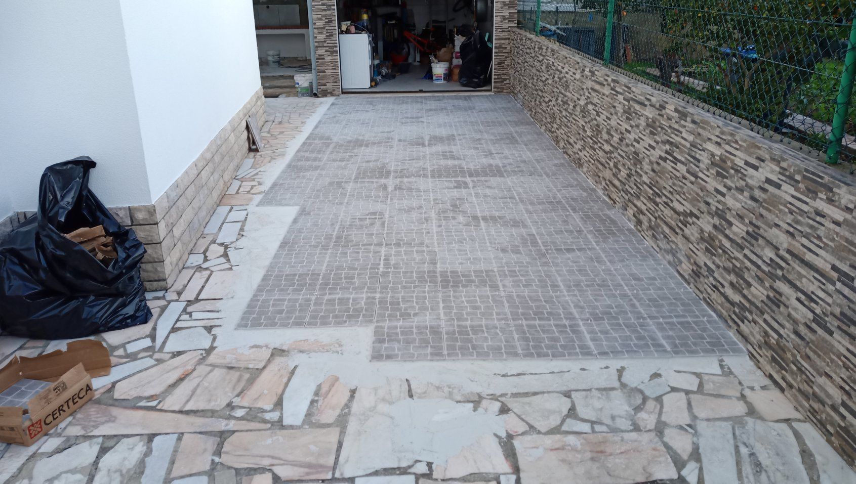 A entrada de veículos é pavimentada com ladrilhos texturizados e borda de pedra, levando a uma garagem. Uma sacola preta está à esquerda.