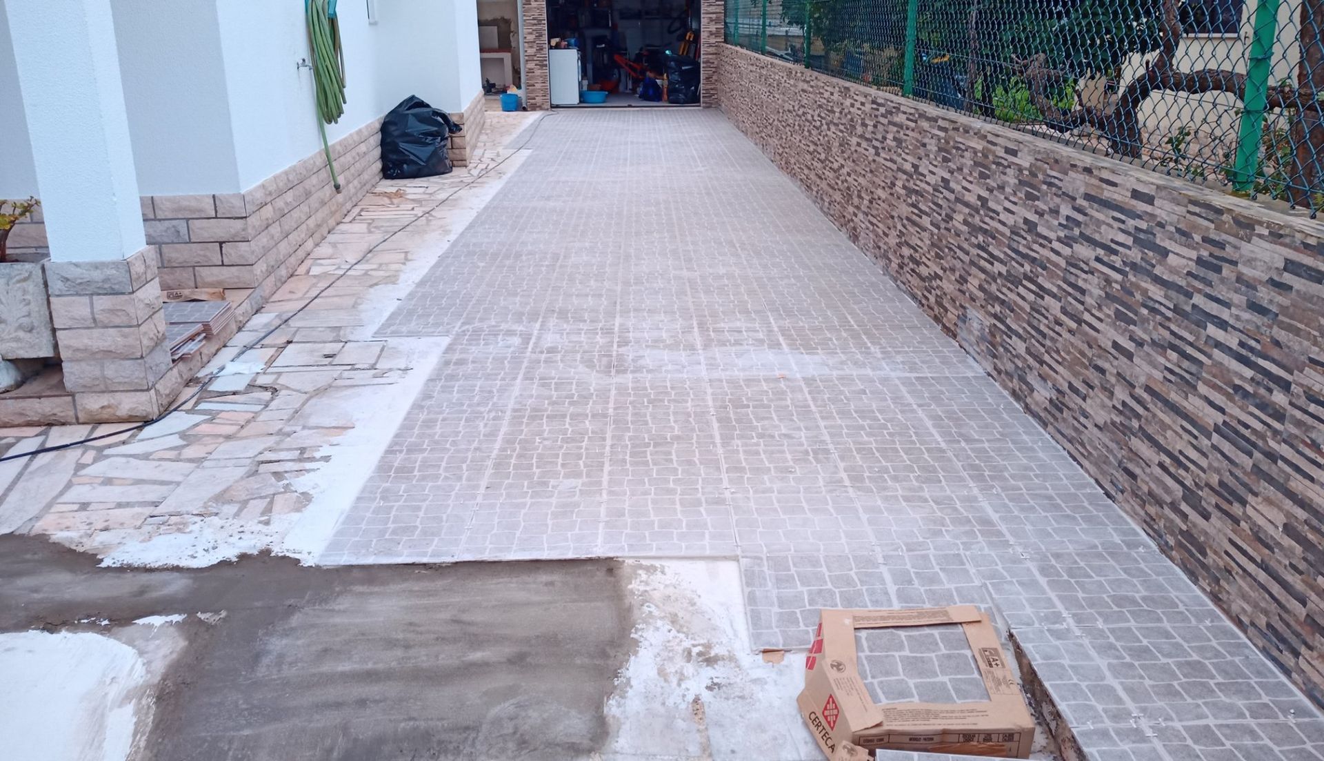Longa entrada de automóveis pavimentada com ladrilhos quadrados, que leva a uma garagem. Muros de pedra e estuque emolduram o caminho.