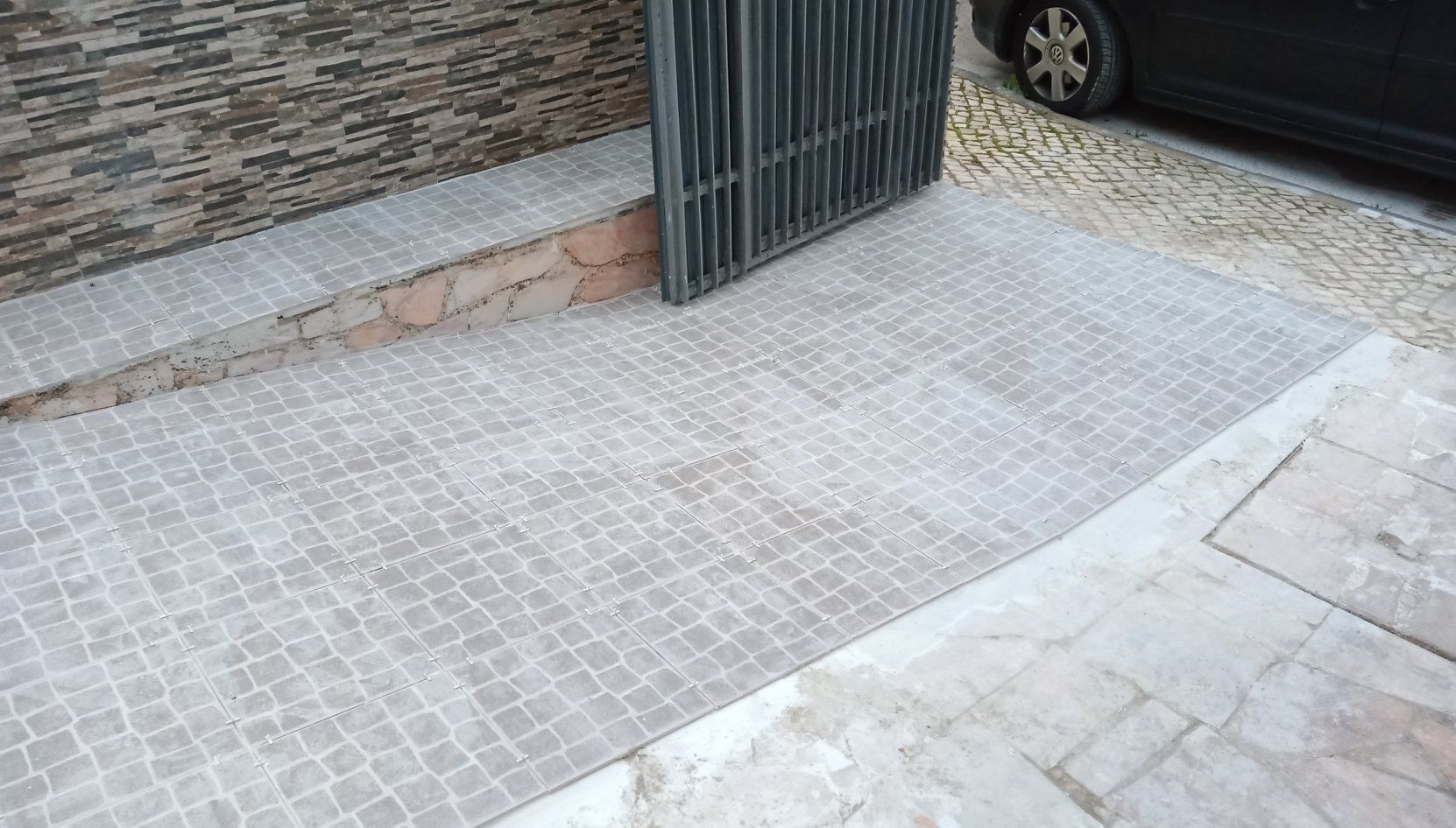 Rampa de acesso a um portão. Superfície de paralelepípedos cinza, pequeno muro à esquerda e pneu de carro visível à direita.