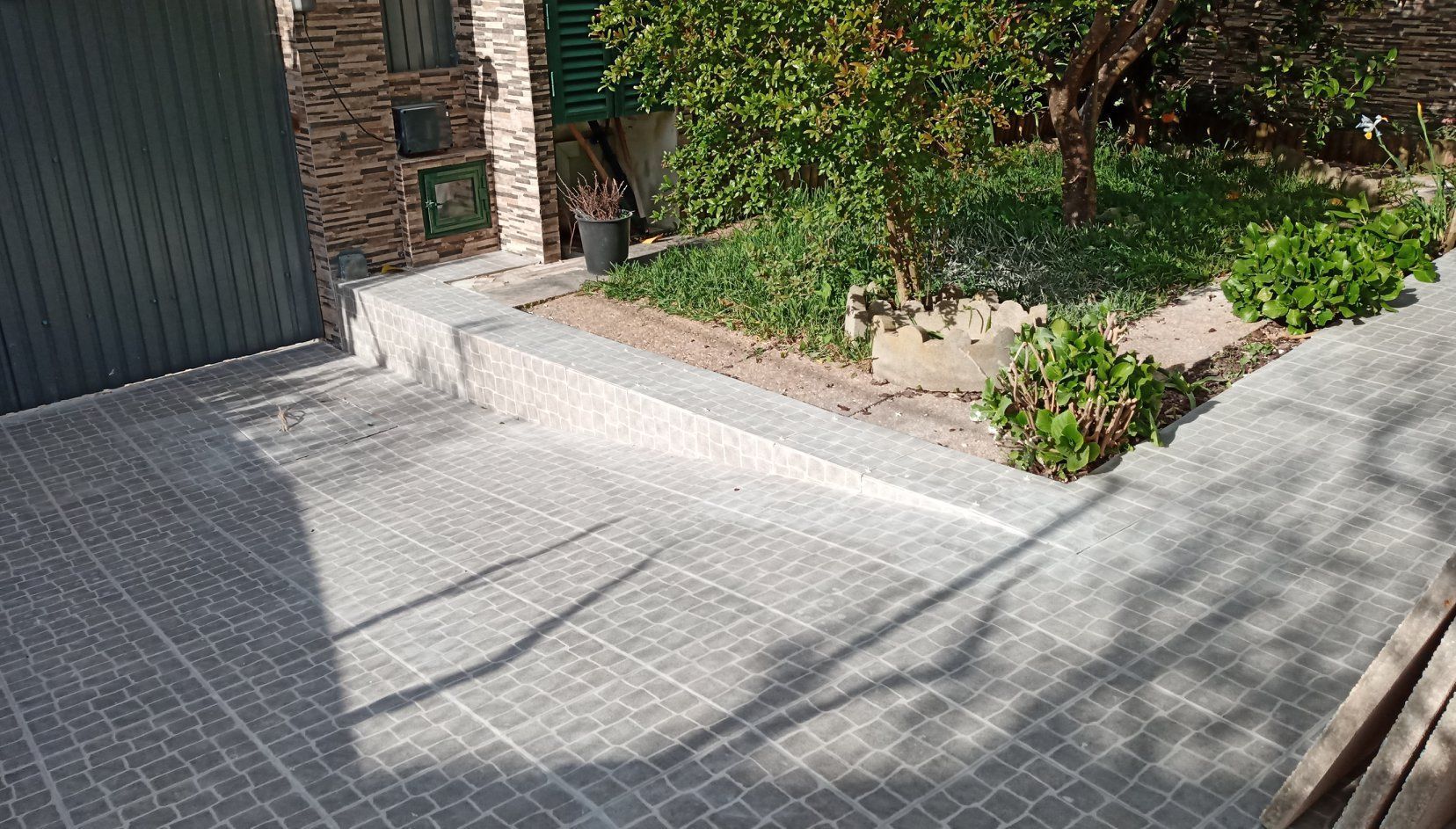 Entrada de veículos e jardim com piso de azulejo e vegetação. Muro com arbustos e árvore.