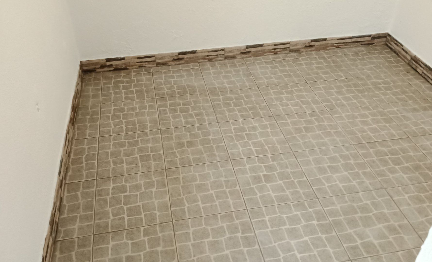 Canto de uma sala com piso de ladrilhos imitando pedra e moldura decorativa. Paredes brancas.