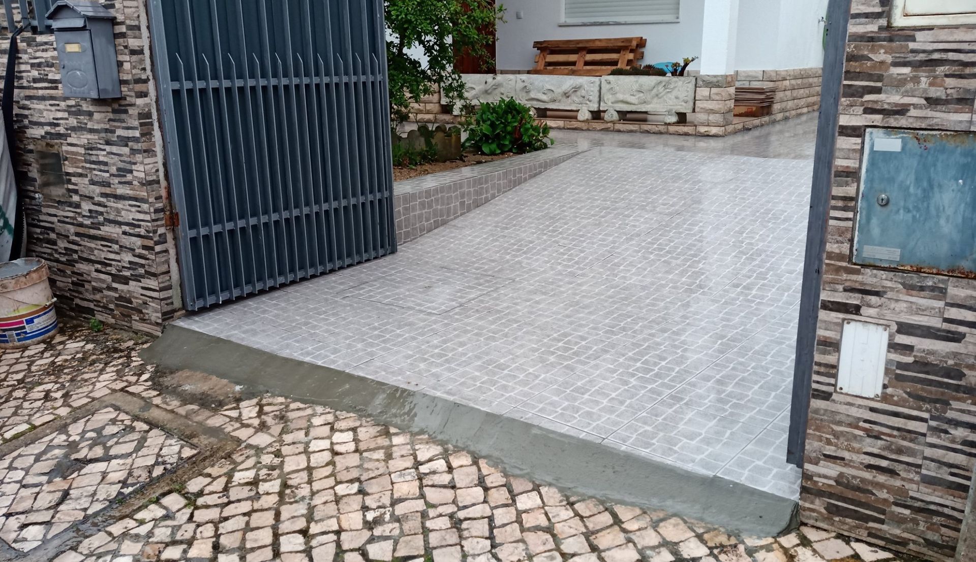 Rampa de entrada com portão que dá acesso a um edifício. Superfície pavimentada, em tons de cinza e bege.