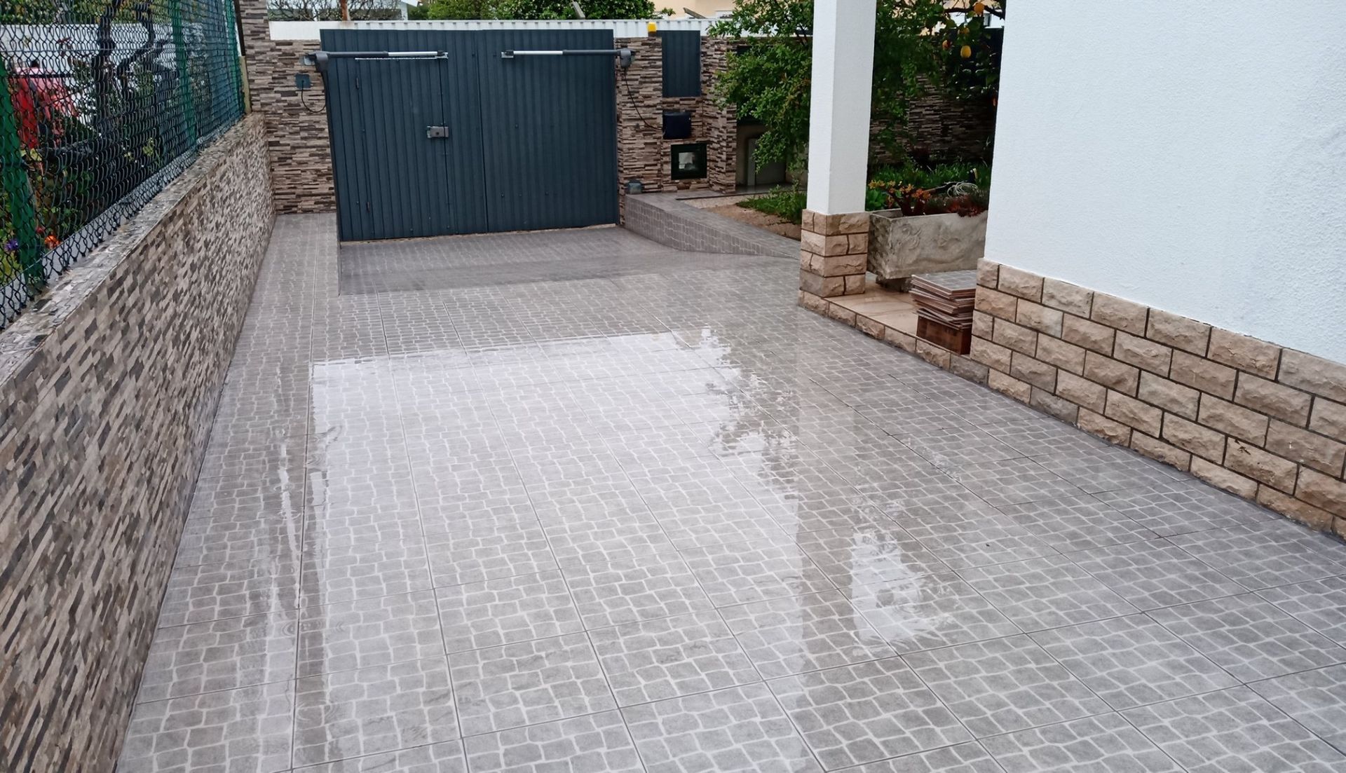 Uma entrada de automóveis pavimentada com pedras molhadas leva a uma porta de garagem cinza. Muro de pedra à esquerda, prédio branco à direita.