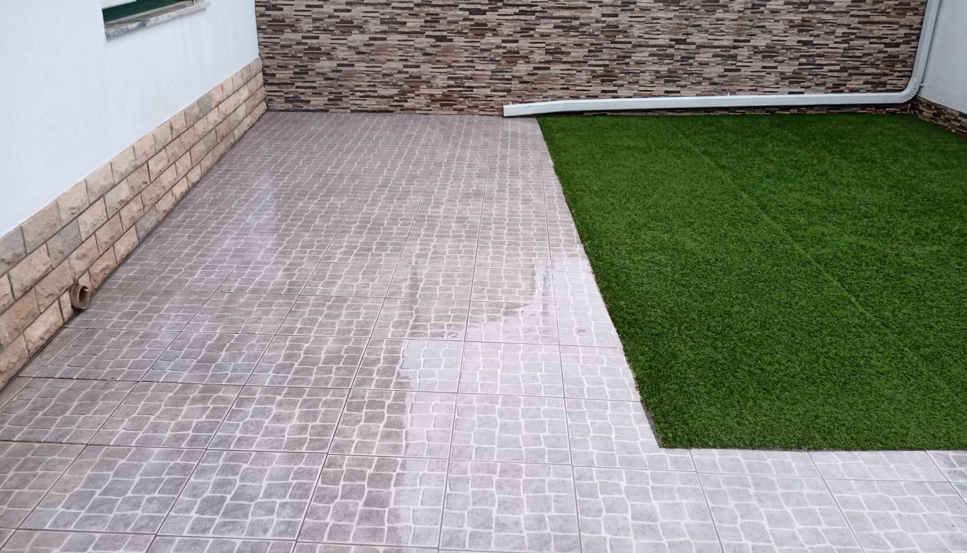 Pátio com ladrilhos de pedra cinza e grama artificial verde. Paredes de pedra e tijolo ao fundo.