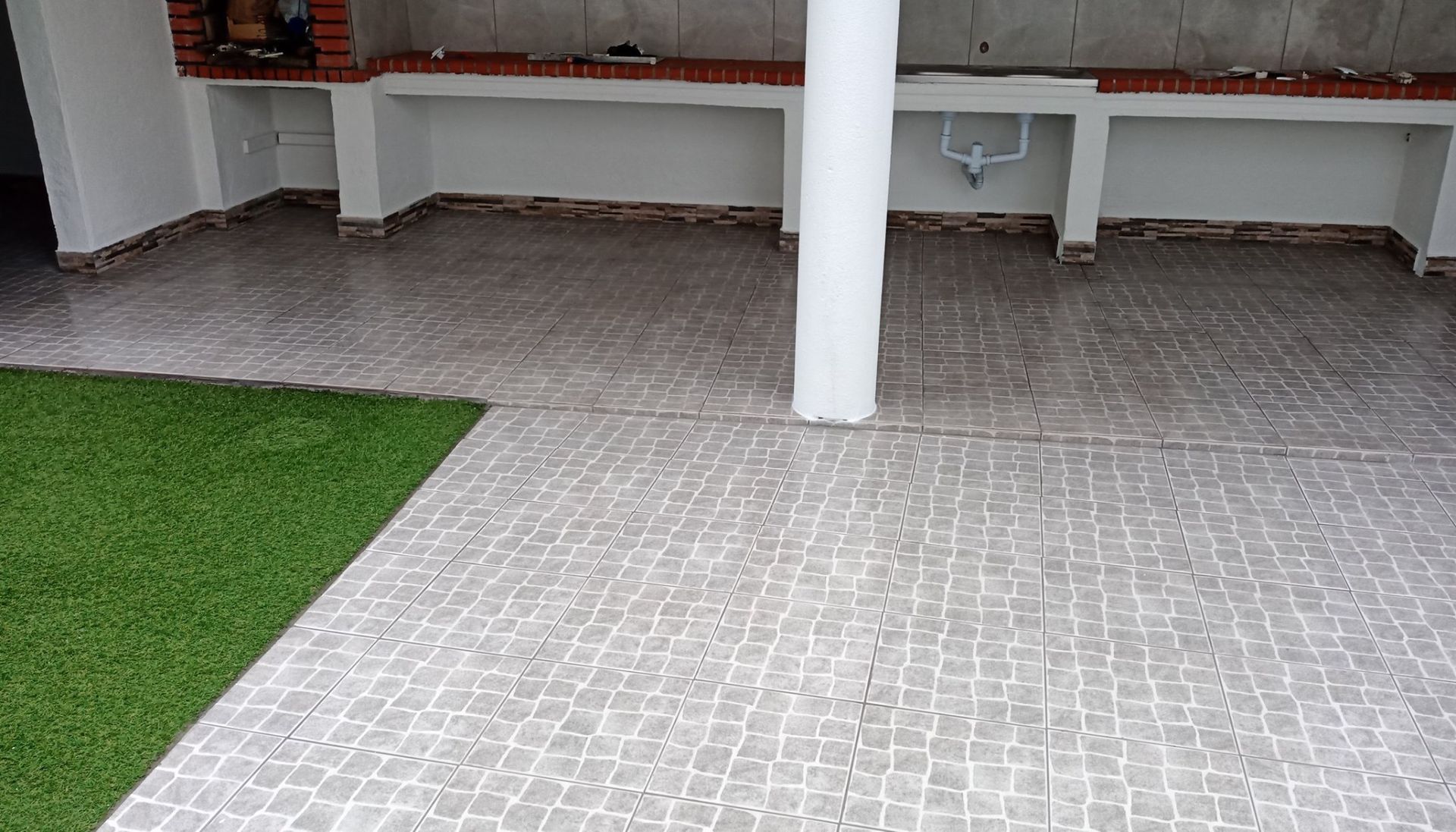 Pátio com piso de azulejos cinza, uma coluna branca e detalhes em tijolo, com grama artificial verde na lateral.