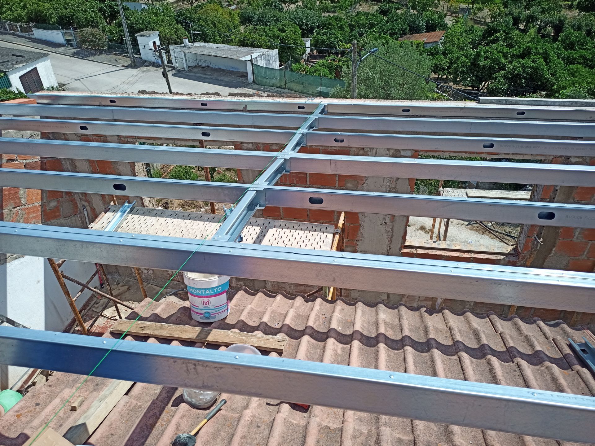 Estrutura metálica instalada em um telhado de telhas vermelhas, provavelmente para um projeto de construção, com um jardim visível ao fundo.