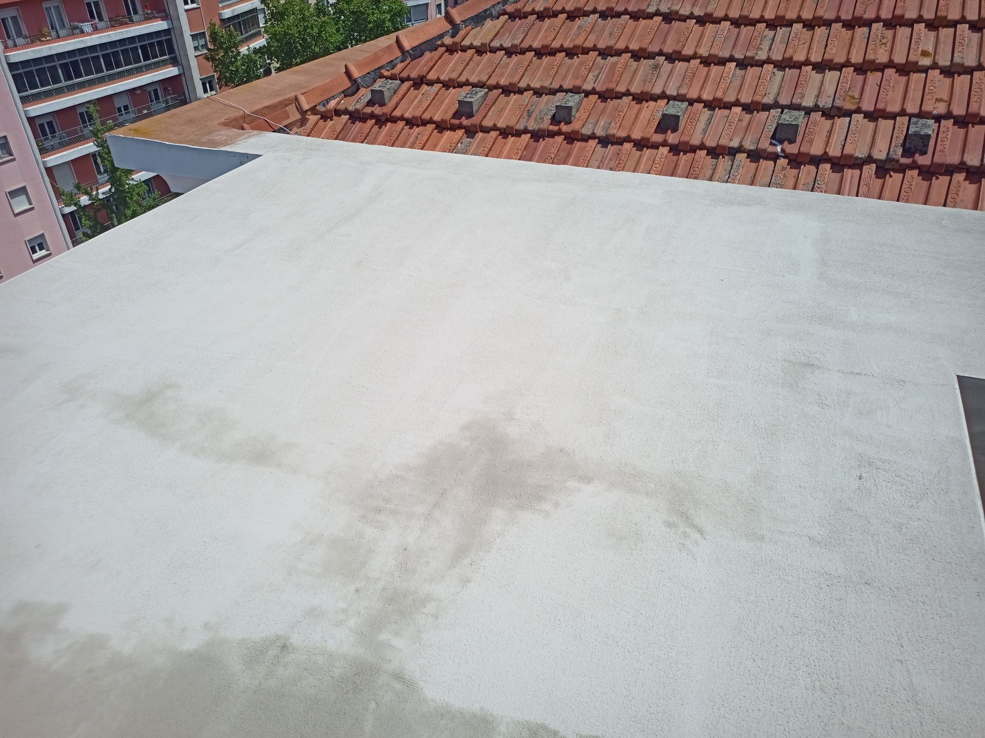 Superfície plana de telhado claro ao lado de um telhado de telhas vermelhas. Prédios e árvores visíveis ao fundo em um dia ensolarado.