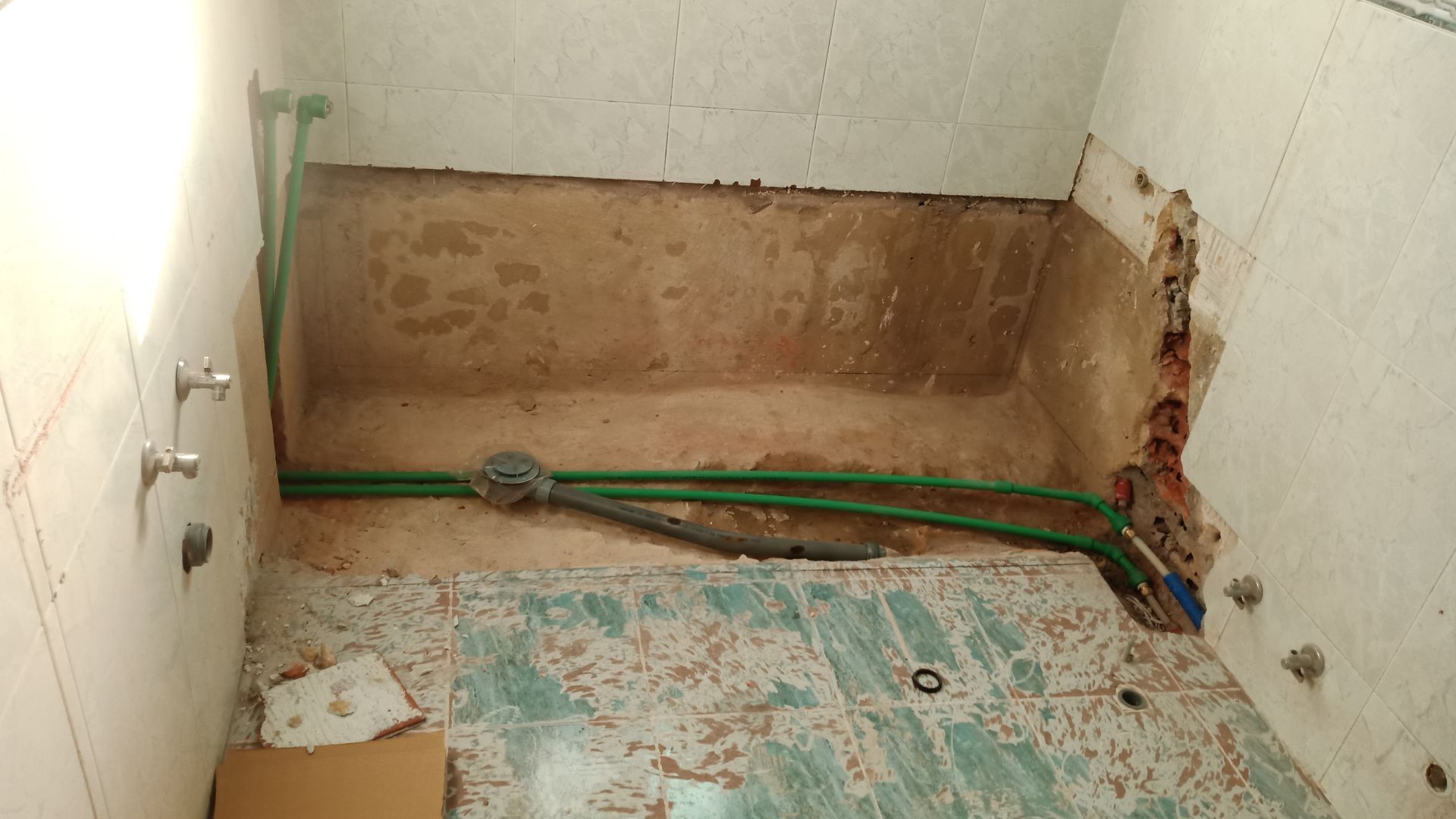 Banheiro reformado com canos expostos e danos na parede. Canos verdes, piso de azulejo azul e branco.