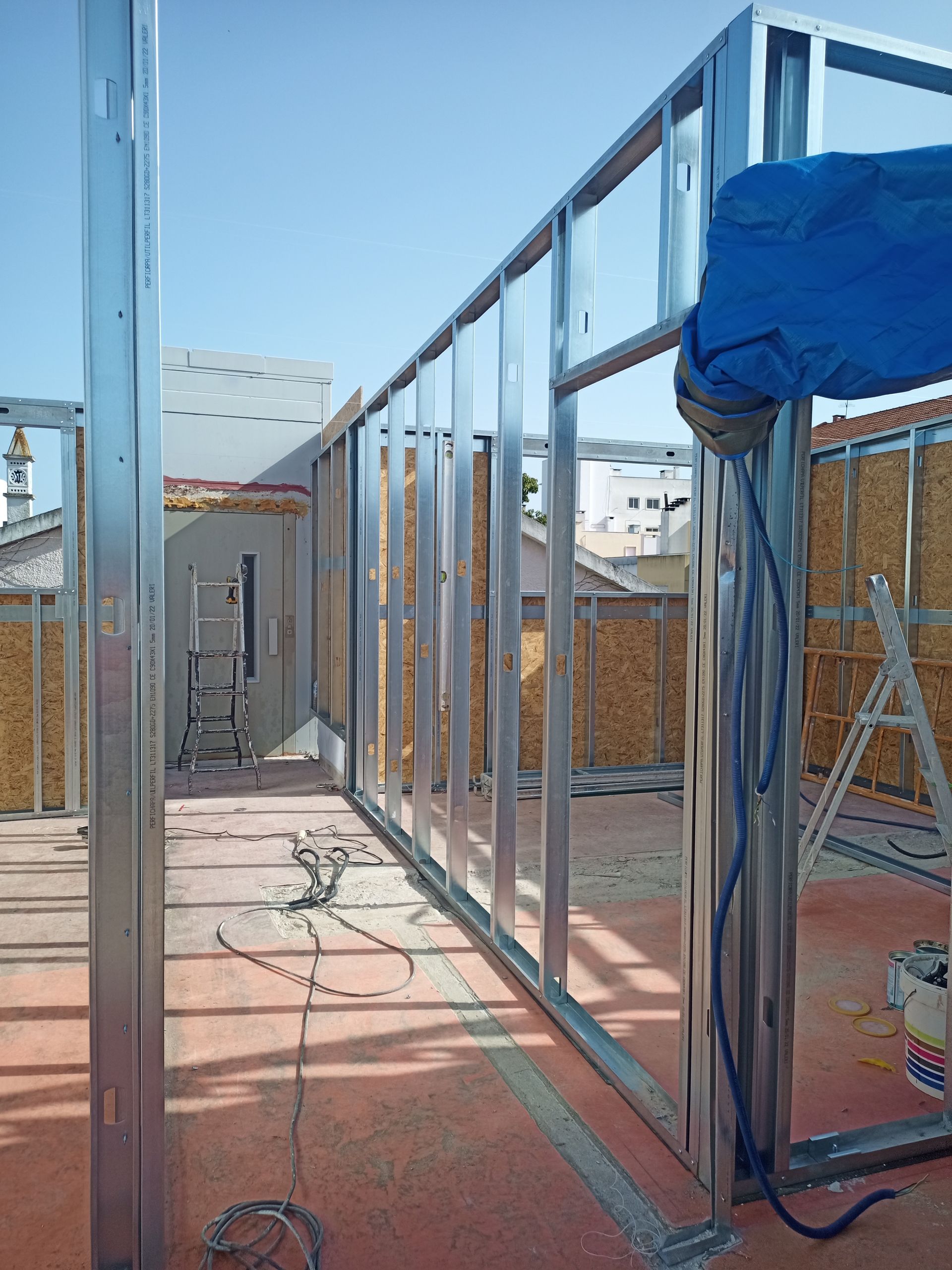 Estrutura metálica de uma parede em construção em um telhado, com uma escada e a lona azul visíveis.