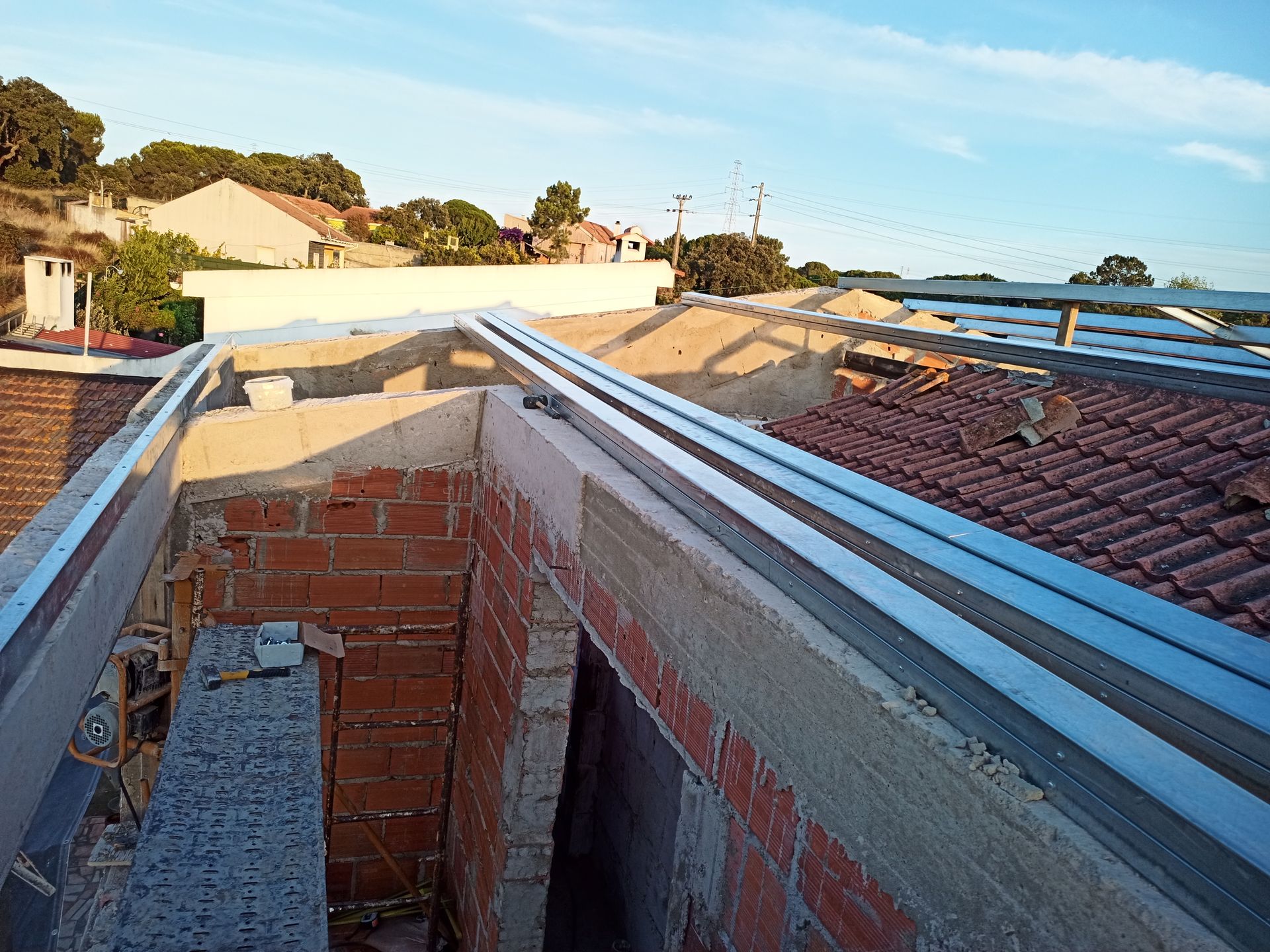Estrutura do telhado: estrutura metálica sobre tijolo e concreto, com casas e árvores ao fundo sob um céu azul.