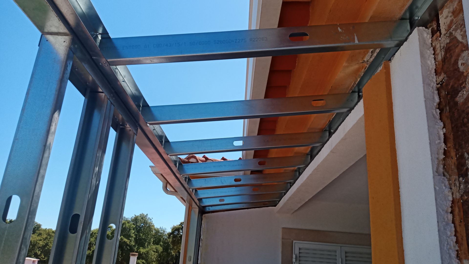 Estrutura metálica para cobertura de varanda contra um céu azul, fixada a uma parede de tijolos e branca.