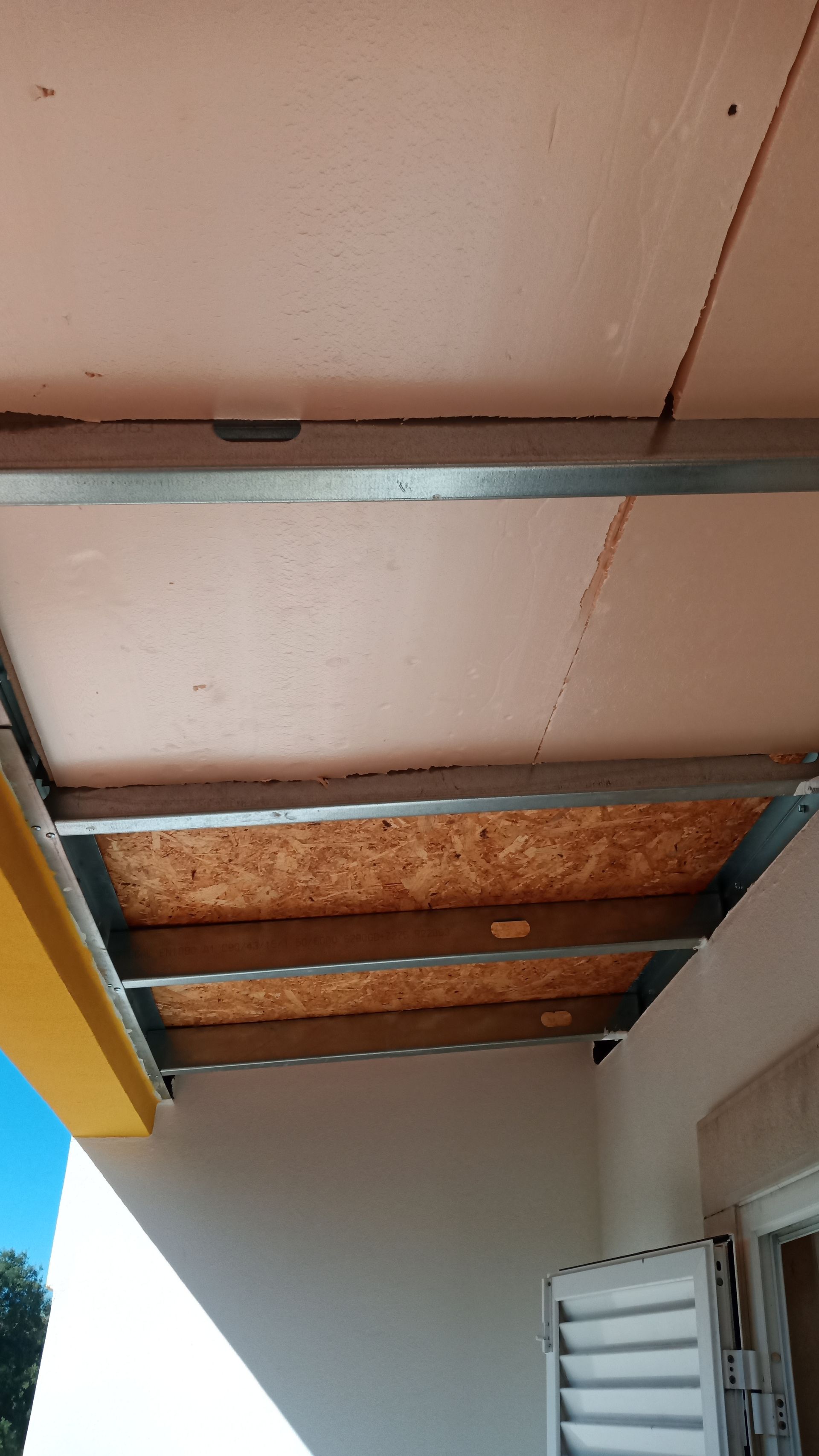 Estrutura do teto: estrutura metálica, isolamento rosa, painéis de madeira marrom. Calha amarela, parede branca.