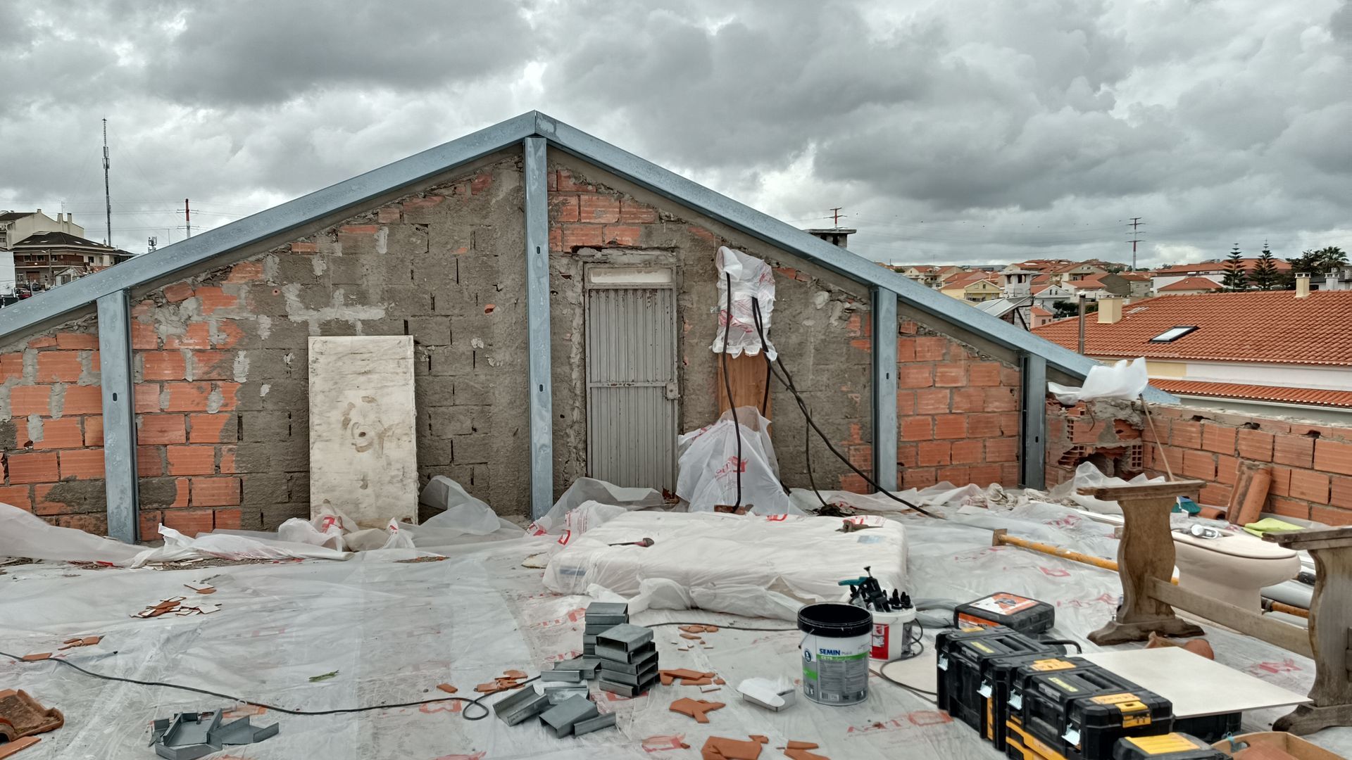 Construção no telhado: parede de tijolos inacabada, estrutura metálica, entulho, céu nublado.