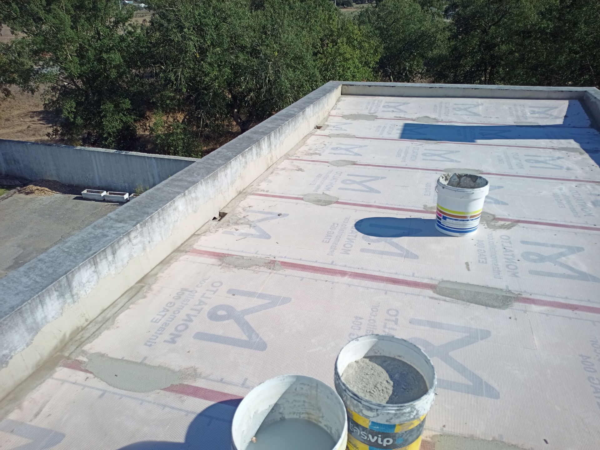 Telhado plano com material rosa e branco, borda de concreto, baldes de material na superfície, árvores ao fundo.