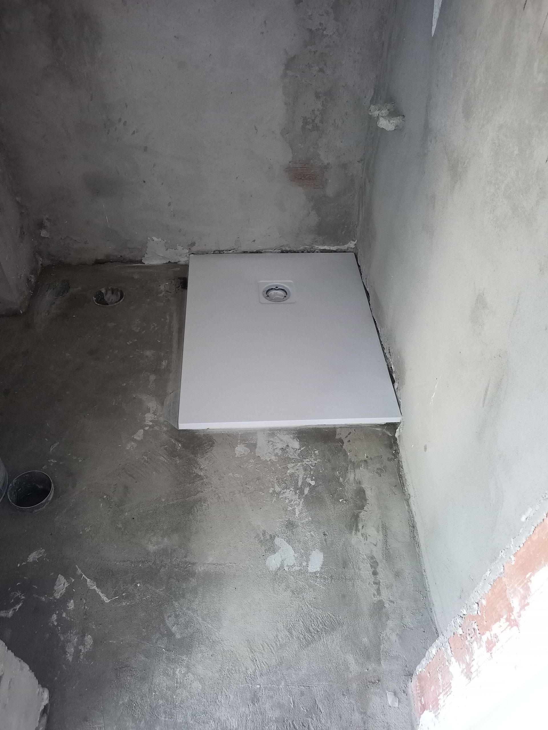 Base de chuveiro retangular branca instalada em um box de chuveiro com paredes de concreto, com um ralo no centro.
