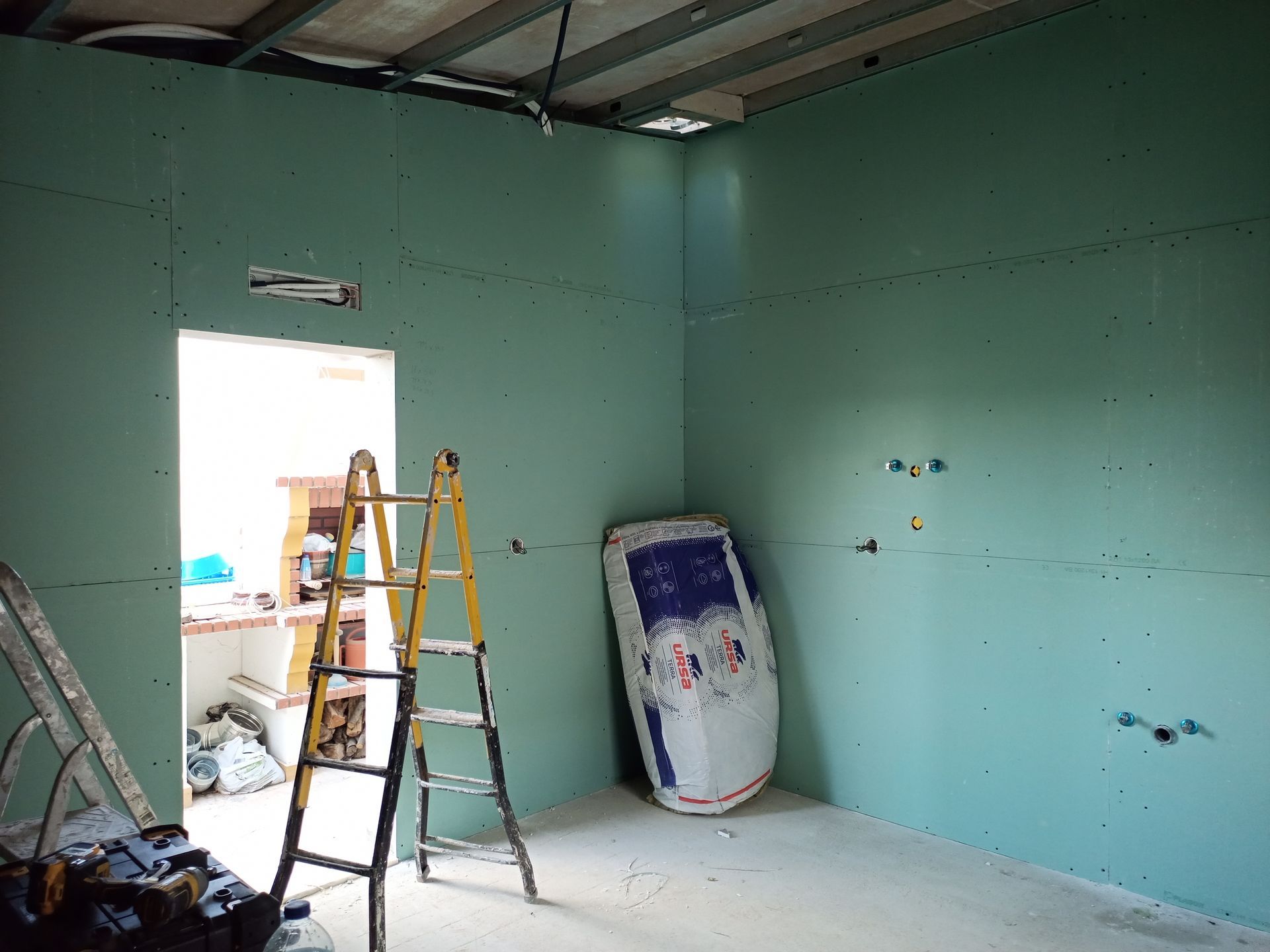 Cômodo em construção com parede de gesso acartonado verde-azulado, uma escada e uma porta se abrindo.