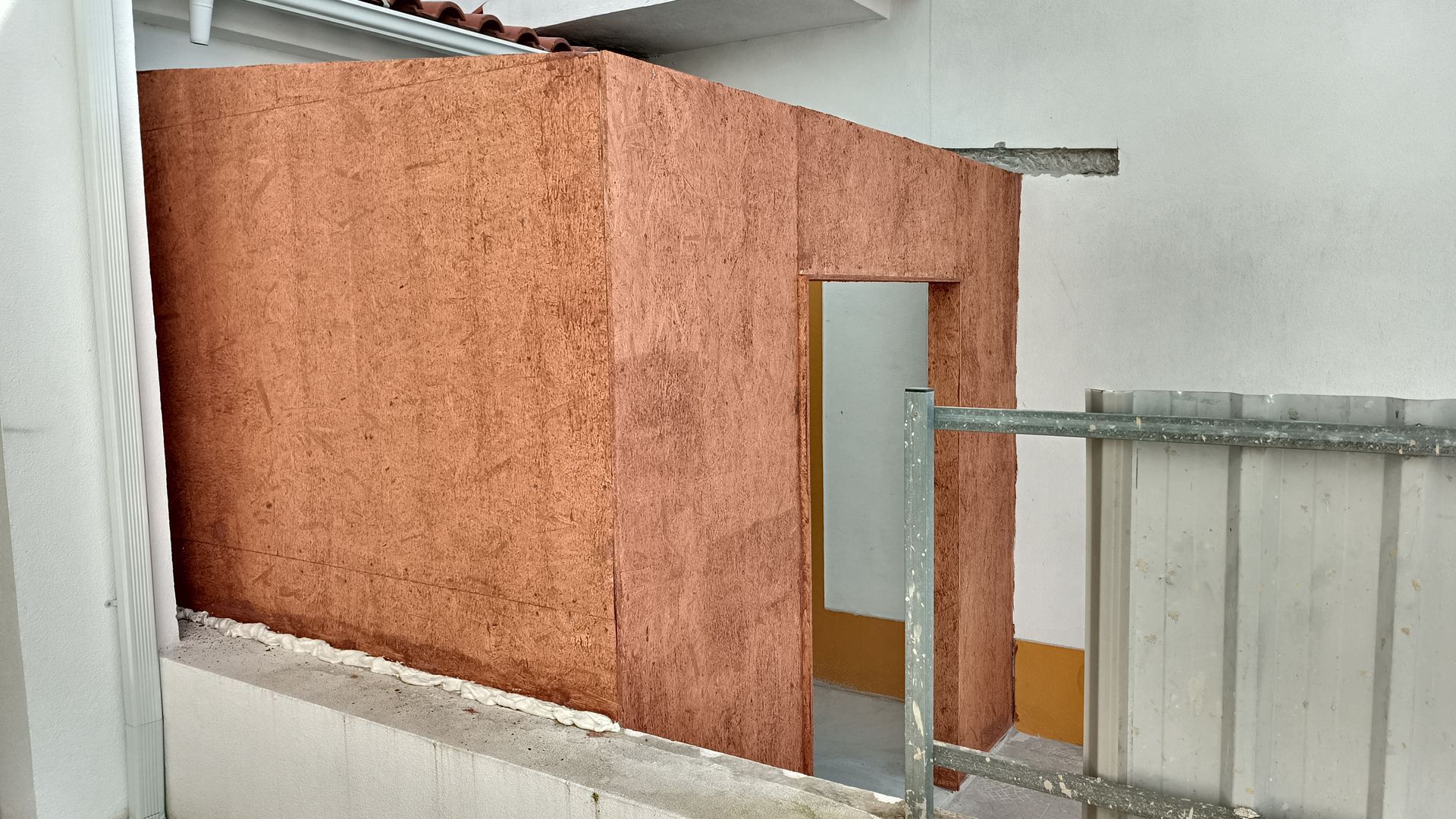 Uma pequena estrutura marrom-avermelhada com uma porta sob um telhado branco. Os suportes de concreto e metal são visíveis.