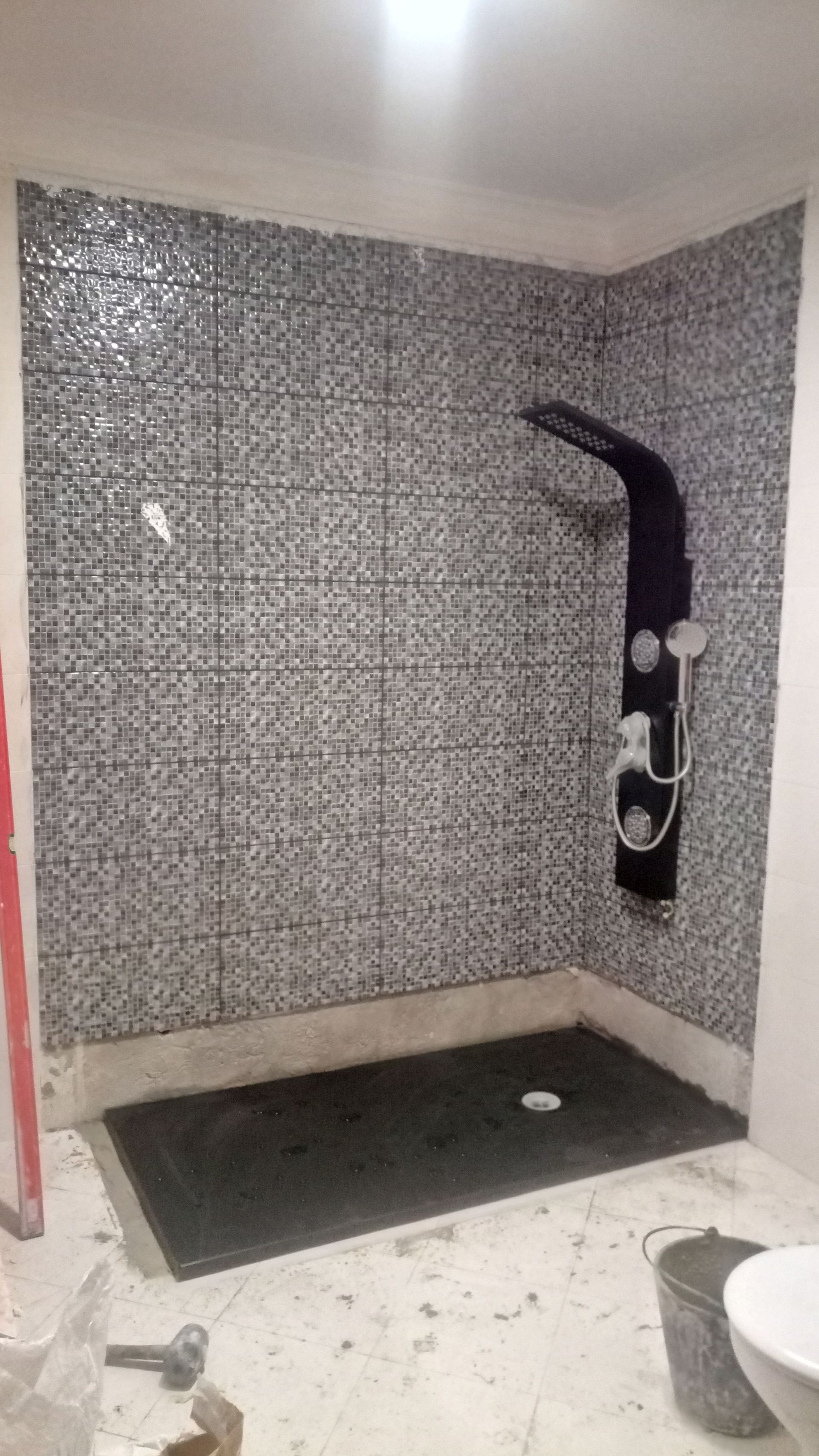 Chuveiro com paredes revestidas de azulejos em mosaico e acessórios pretos. Piso cinza escuro.