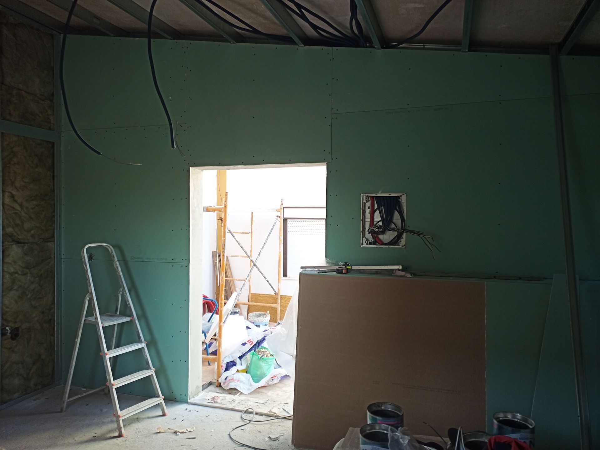 Interior de um cômodo em construção com paredes de gesso acartonado verdes, uma porta aberta e uma escada de mão.