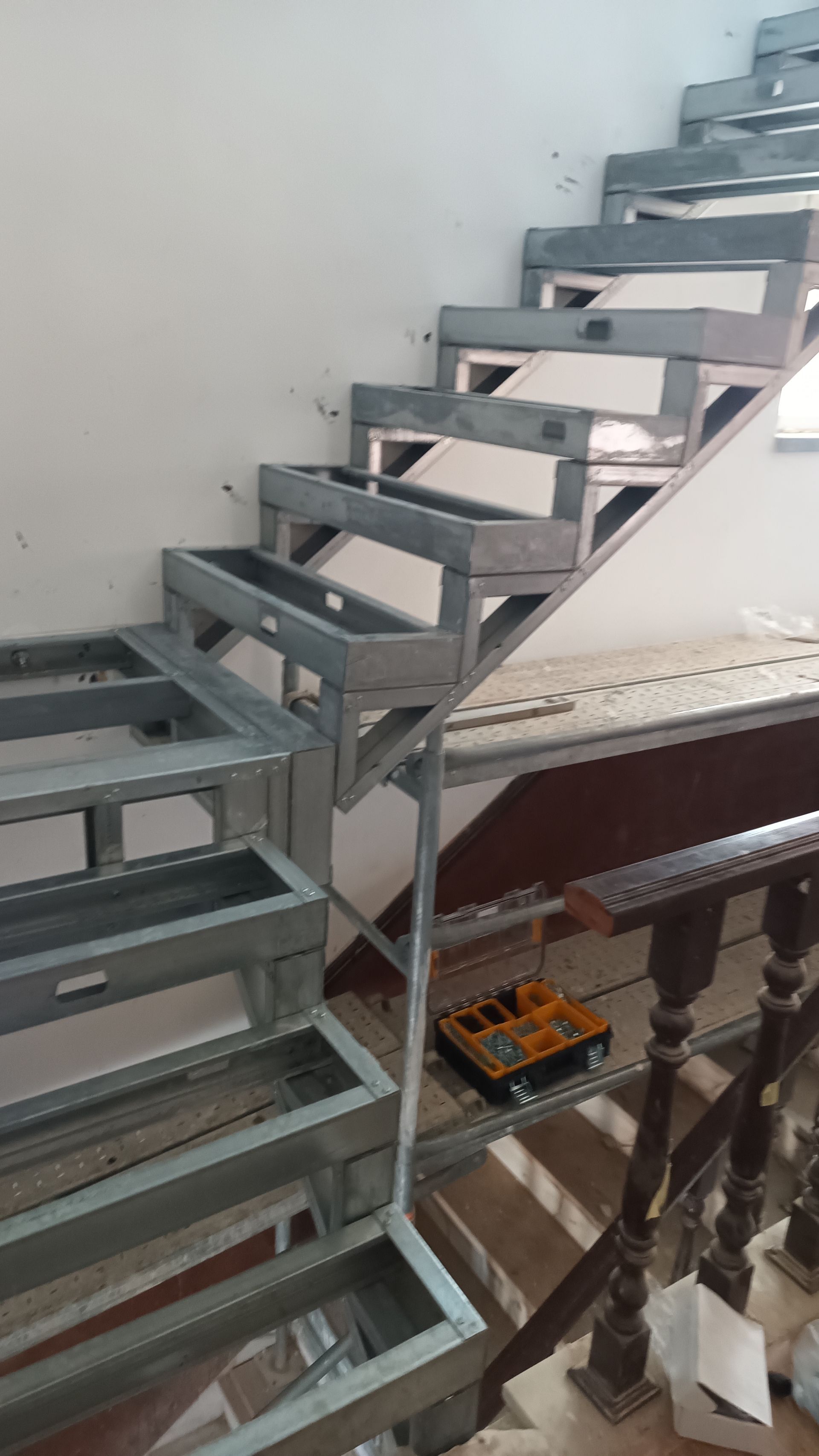 Escada metálica em construção, com degraus vazados e corrimão de madeira.