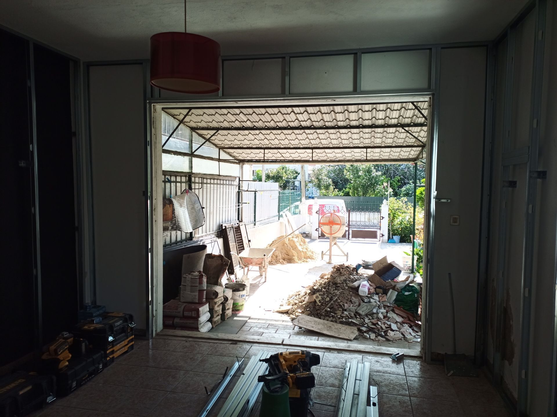 Vista interior de um cômodo em construção, com porta aberta que dá para uma área externa desorganizada com uma garagem coberta.