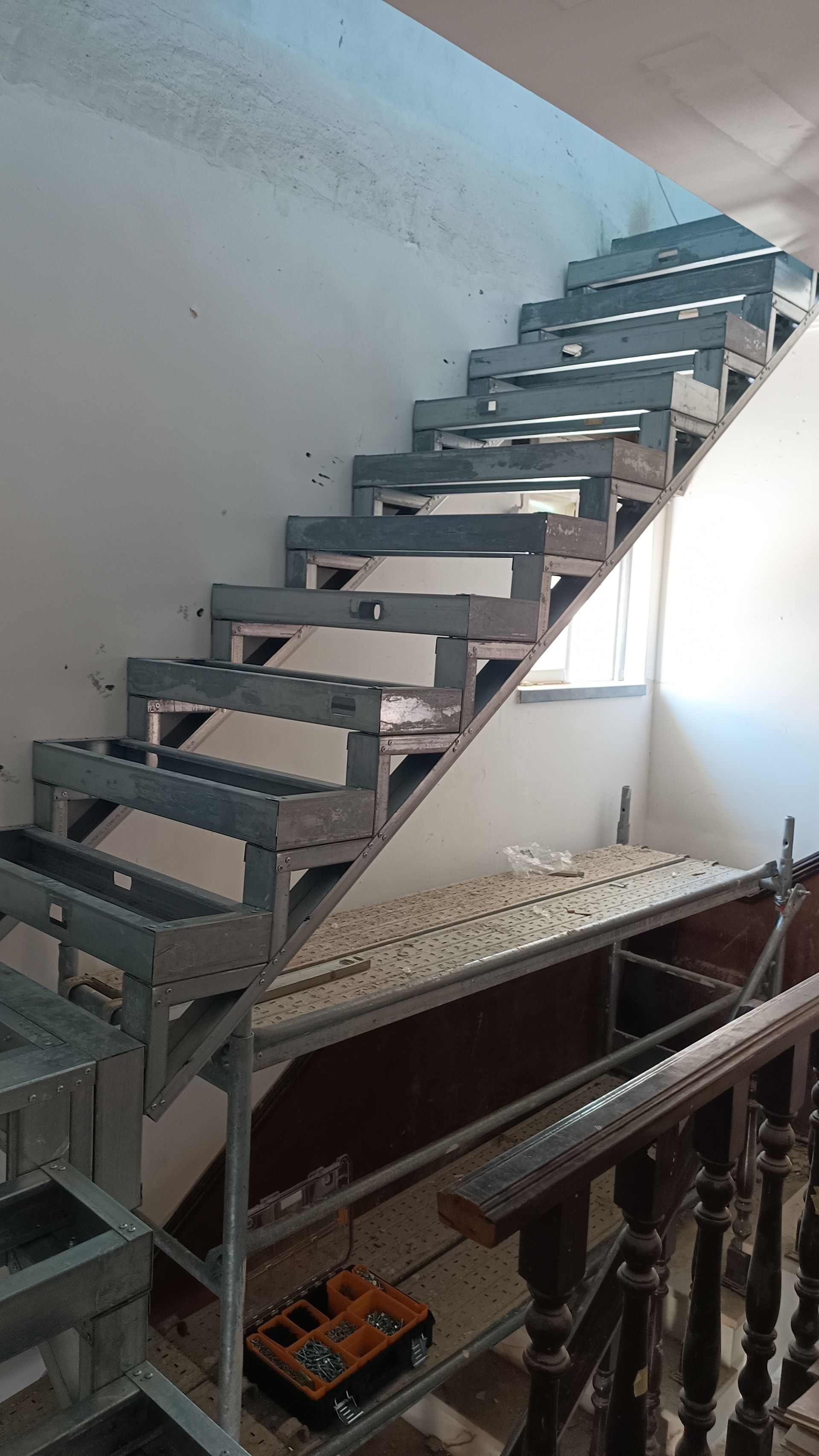 Escada metálica em construção, com espelhos abertos e corrimão visível no nível inferior.