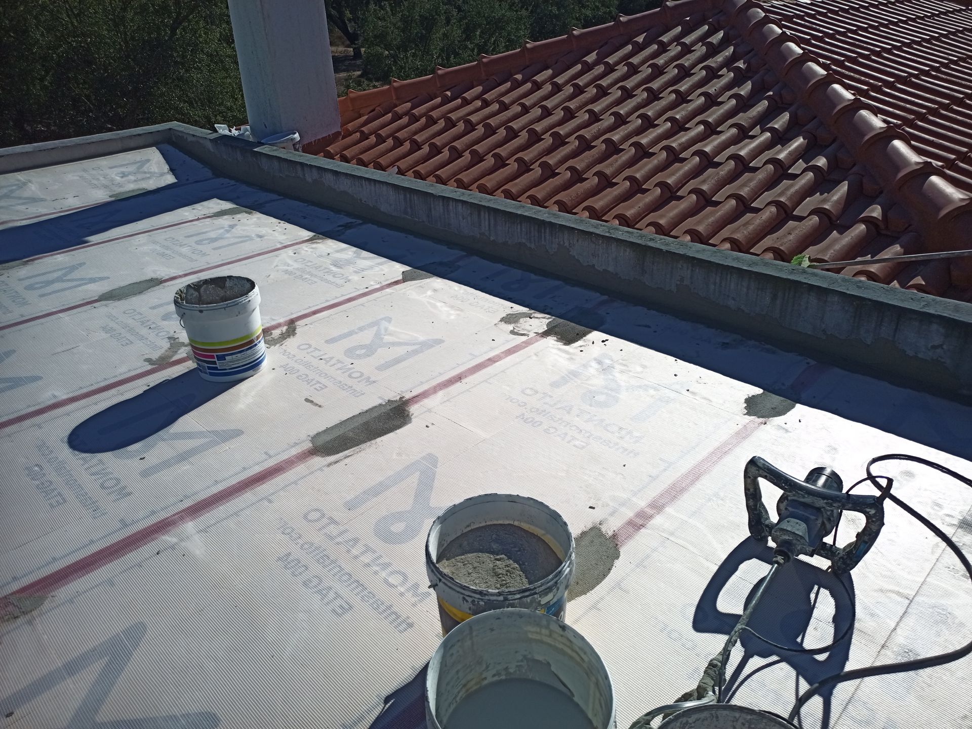Construção de telhado plano em andamento; baldes, betoneira e material na superfície, com um telhado de telhas ao fundo.
