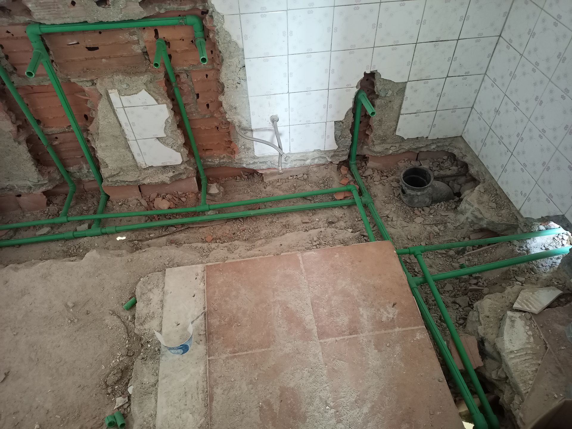 Tubulações de encanamento verdes instaladas em um cômodo com paredes de tijolos aparentes, azulejos e piso em reforma.