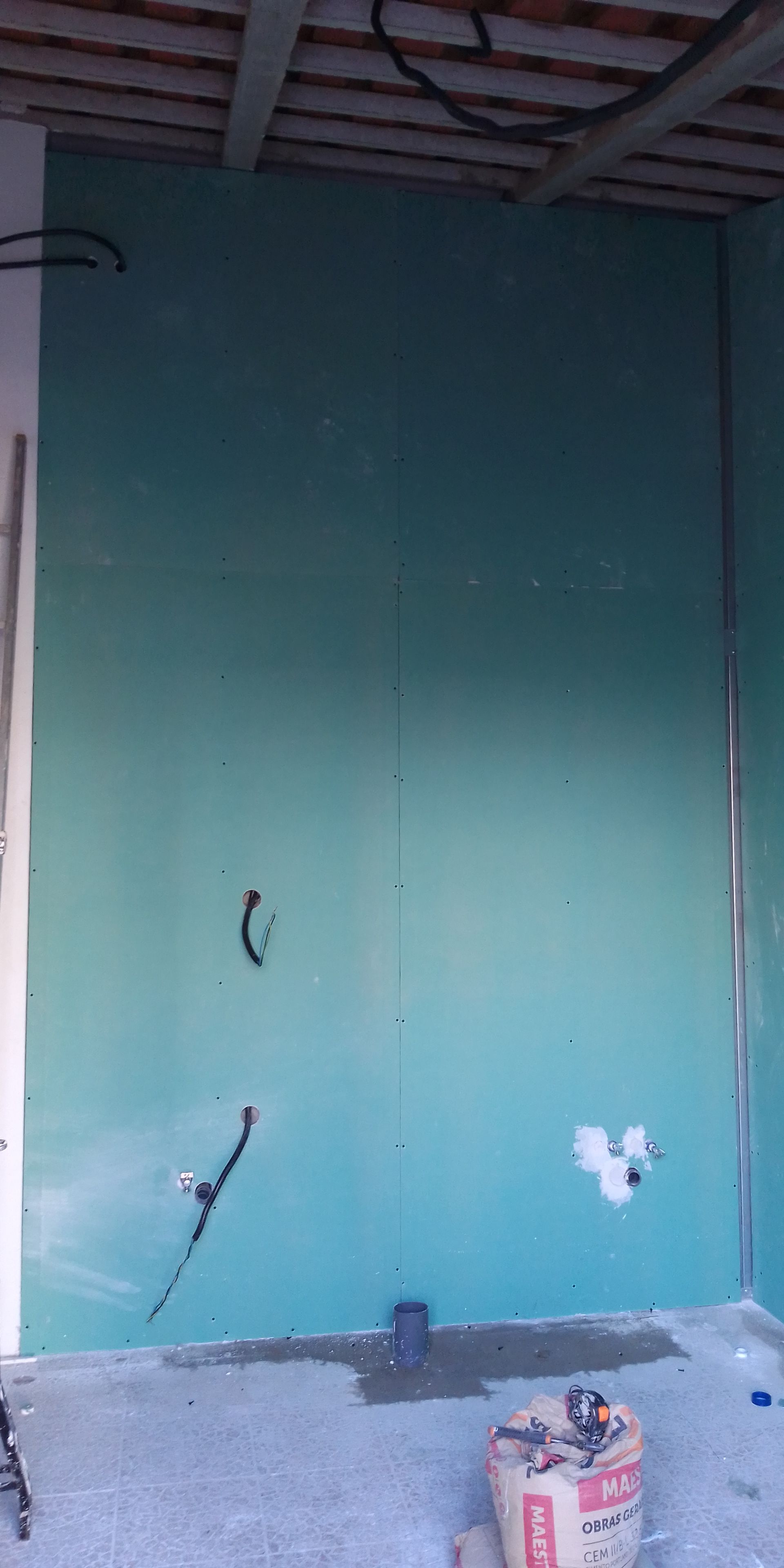 Parede interna em construção com drywall na cor verde-azulada. Fiação e aberturas elétricas visíveis; piso de concreto.