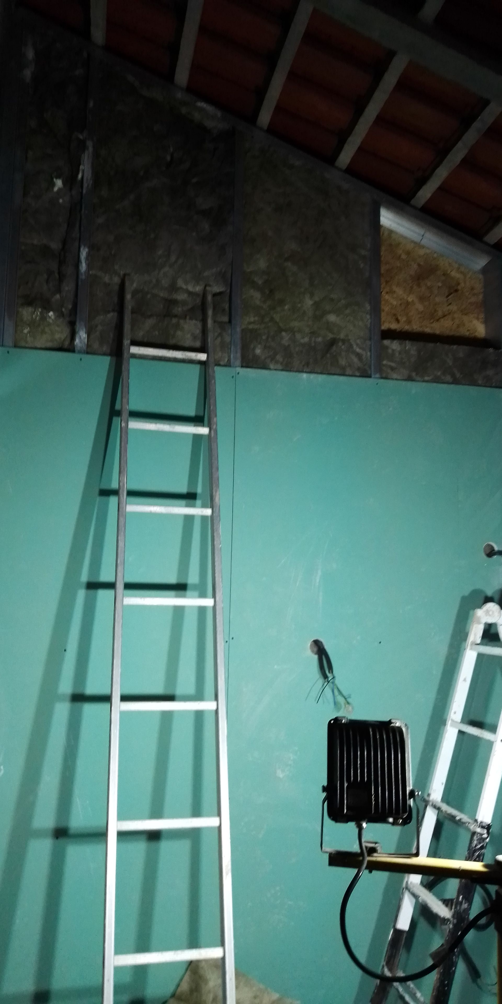 Uma escada de metal está encostada em uma parede verde-azulada. O telhado tem vigas de madeira expostas e telhas vermelhas. Outra escada está perto de um objeto preto.