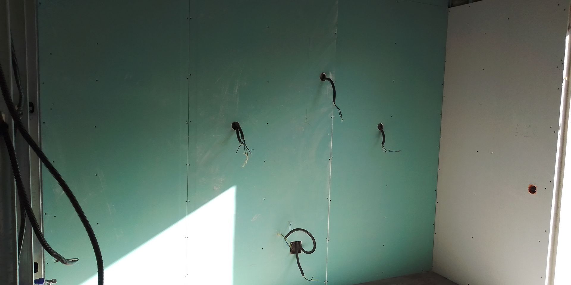 Construção de parede interna com fios elétricos expostos emergindo do drywall verde.