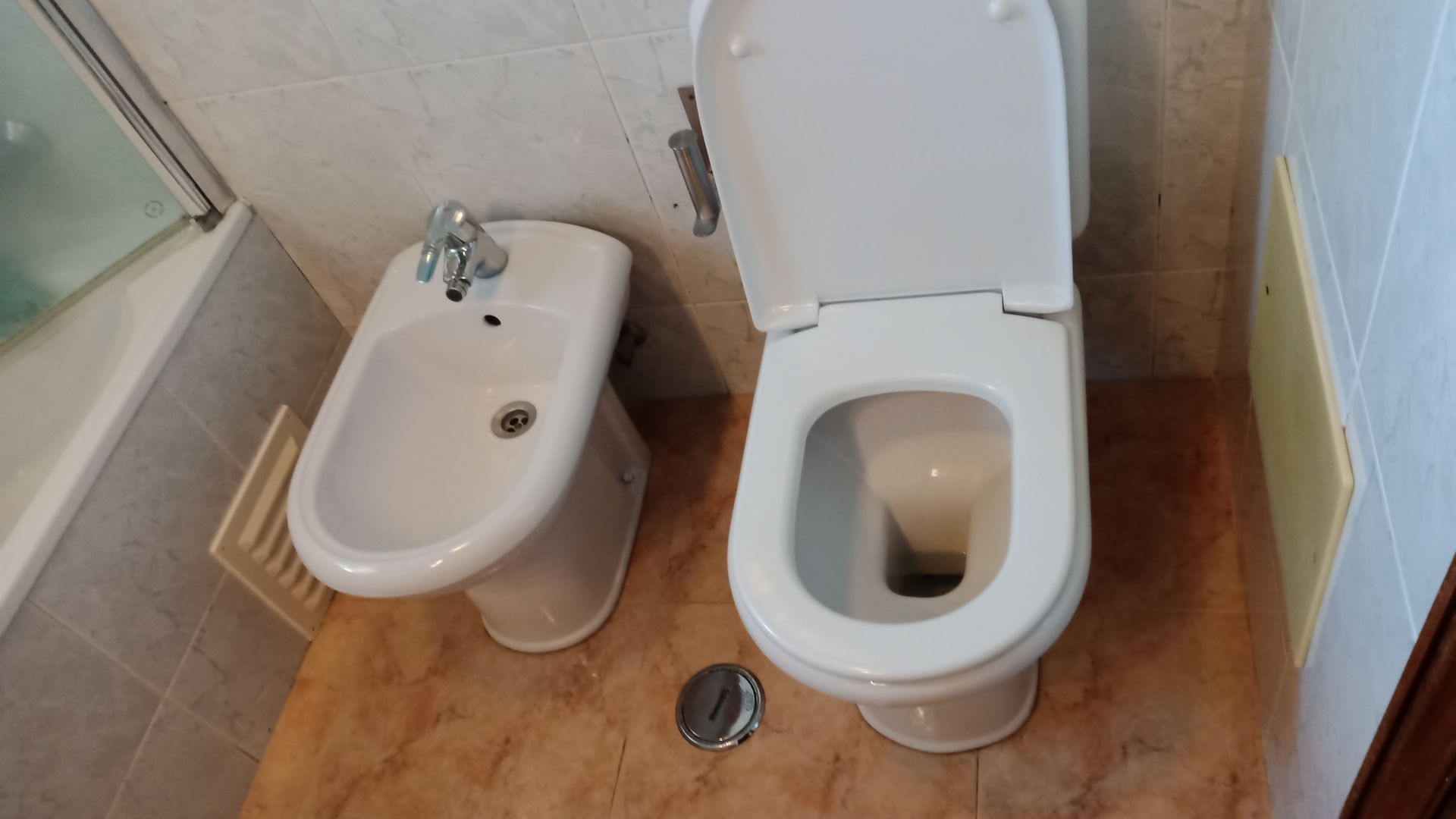 Vaso sanitário e bidê lado a lado em um banheiro. Louças brancas sobre piso de azulejo marrom.