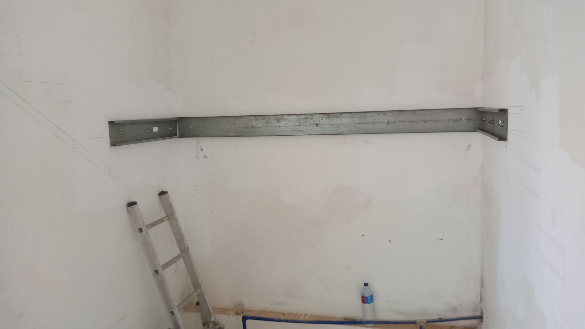 Suporte de prateleira de metal fixado em uma parede branca em um cômodo; uma escada está encostada na parede.