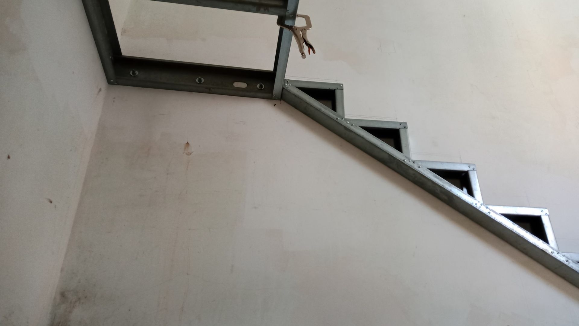 Estrutura metálica da escada embutida em uma parede branca, que leva a uma abertura no teto.