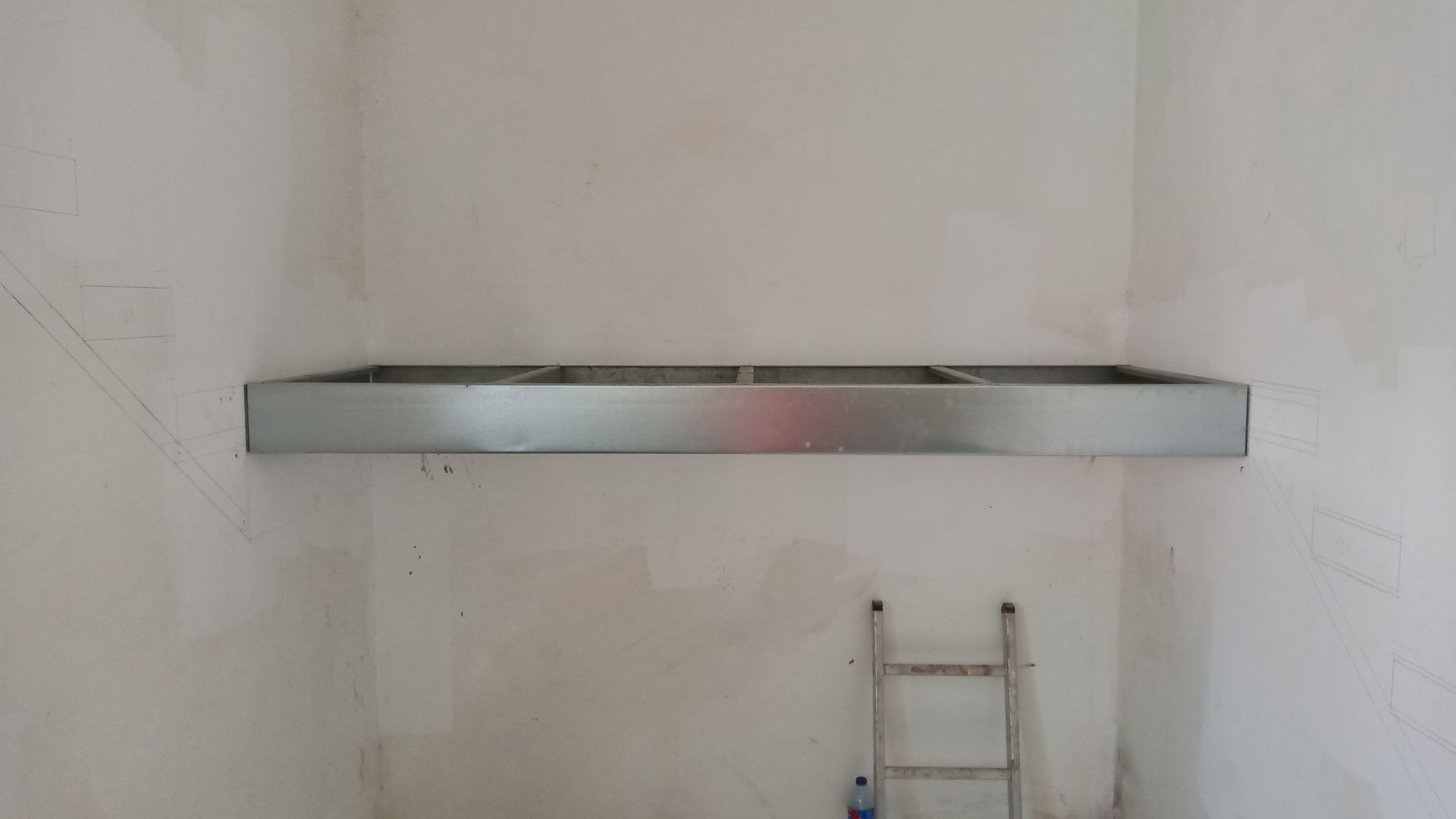 Estrutura de prateleira metálica fixada em uma parede branca em um cômodo em construção, com uma escada ao fundo.