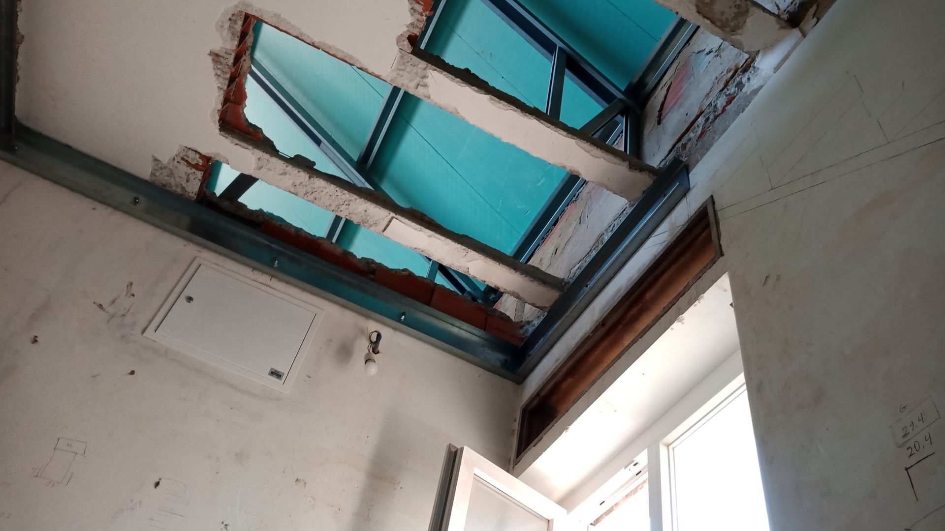 Teto danificado com vigas expostas e céu à mostra, detritos no chão e uma janela em um quarto branco.
