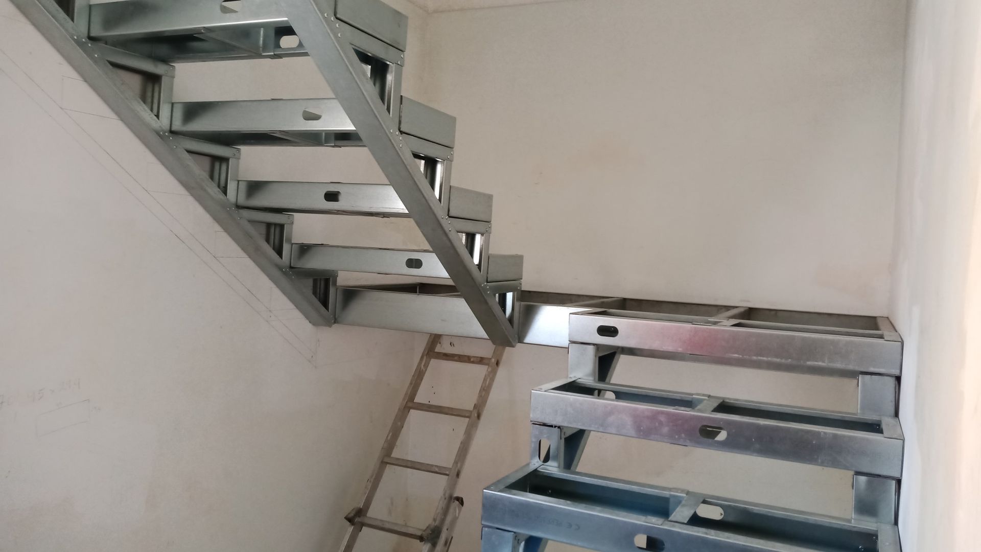 Estrutura metálica de escada instalada em um cômodo com escada de mão e paredes inacabadas.