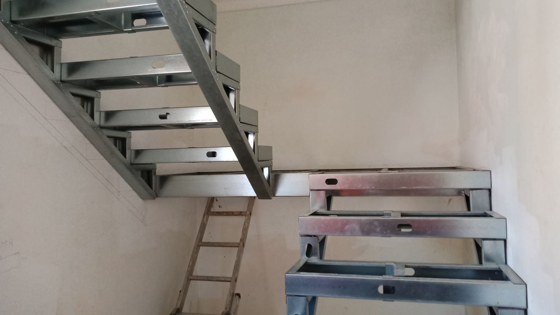 Estrutura metálica de escada em construção dentro de uma sala com uma escada de mão próxima.