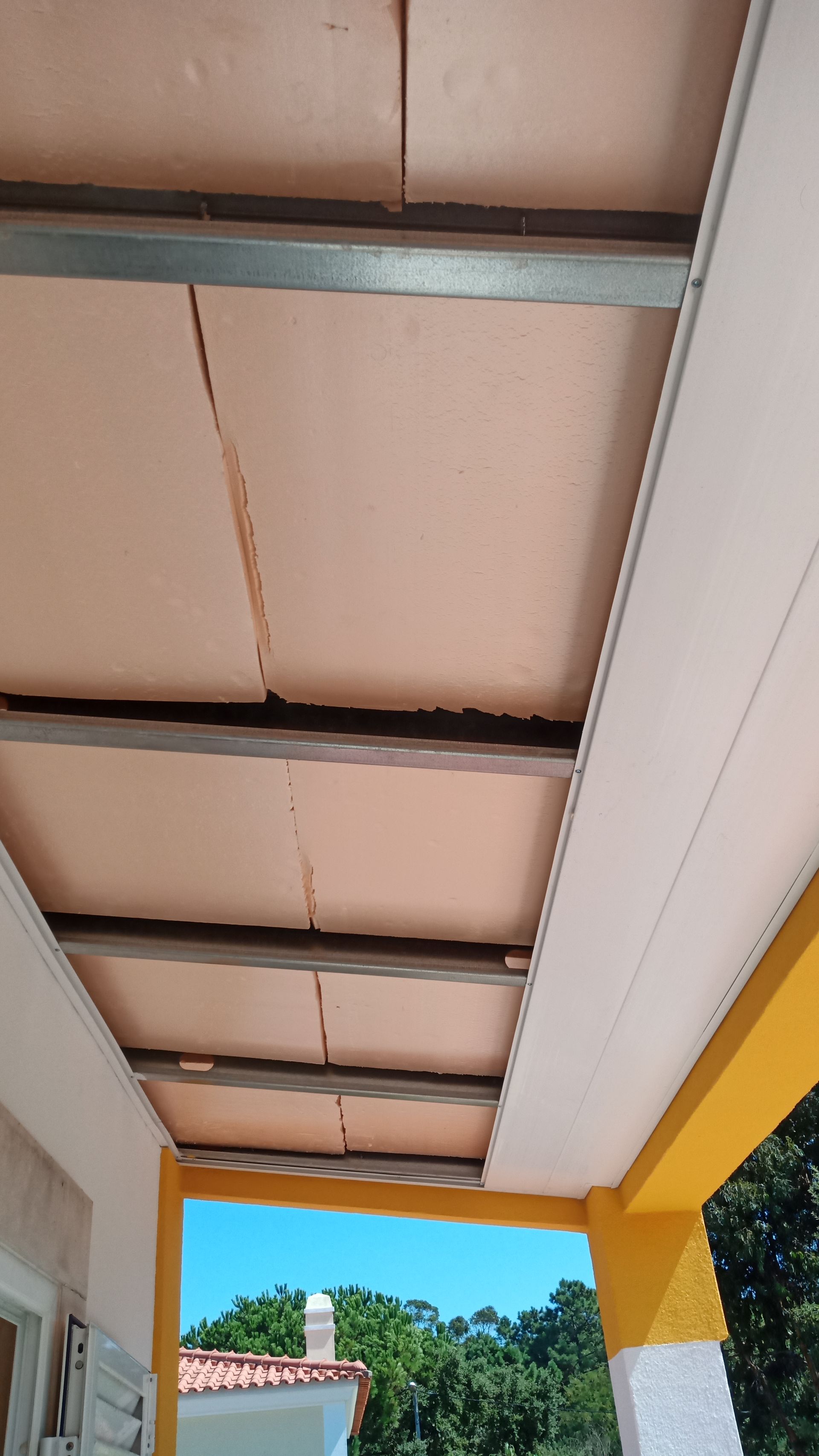 Painéis de teto bege instalados em estrutura metálica, vistos de baixo. Detalhes em amarelo e branco, céu azul ao fundo.