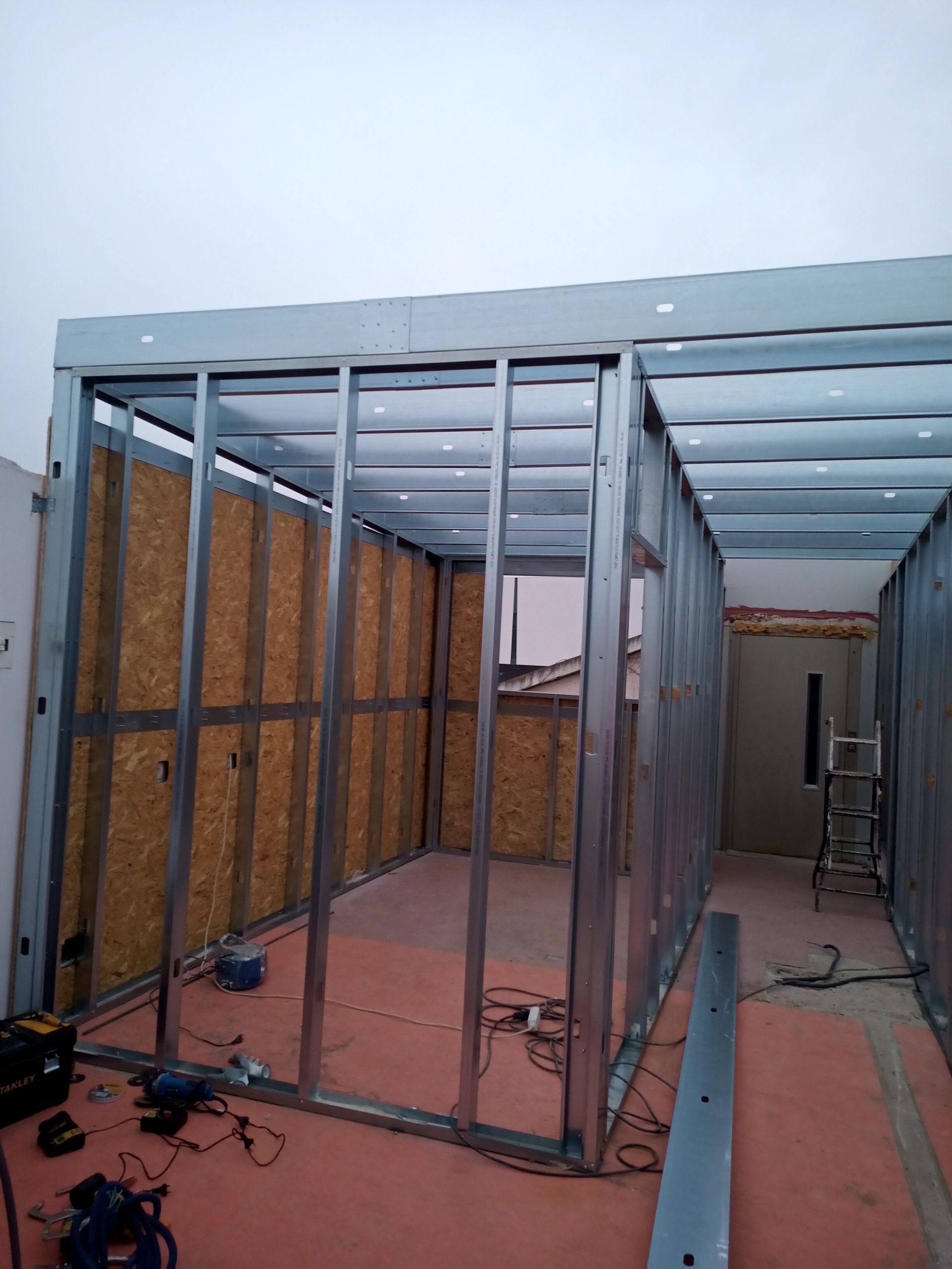 Estrutura metálica de uma sala em construção, com painéis de madeira e ferramentas no chão.