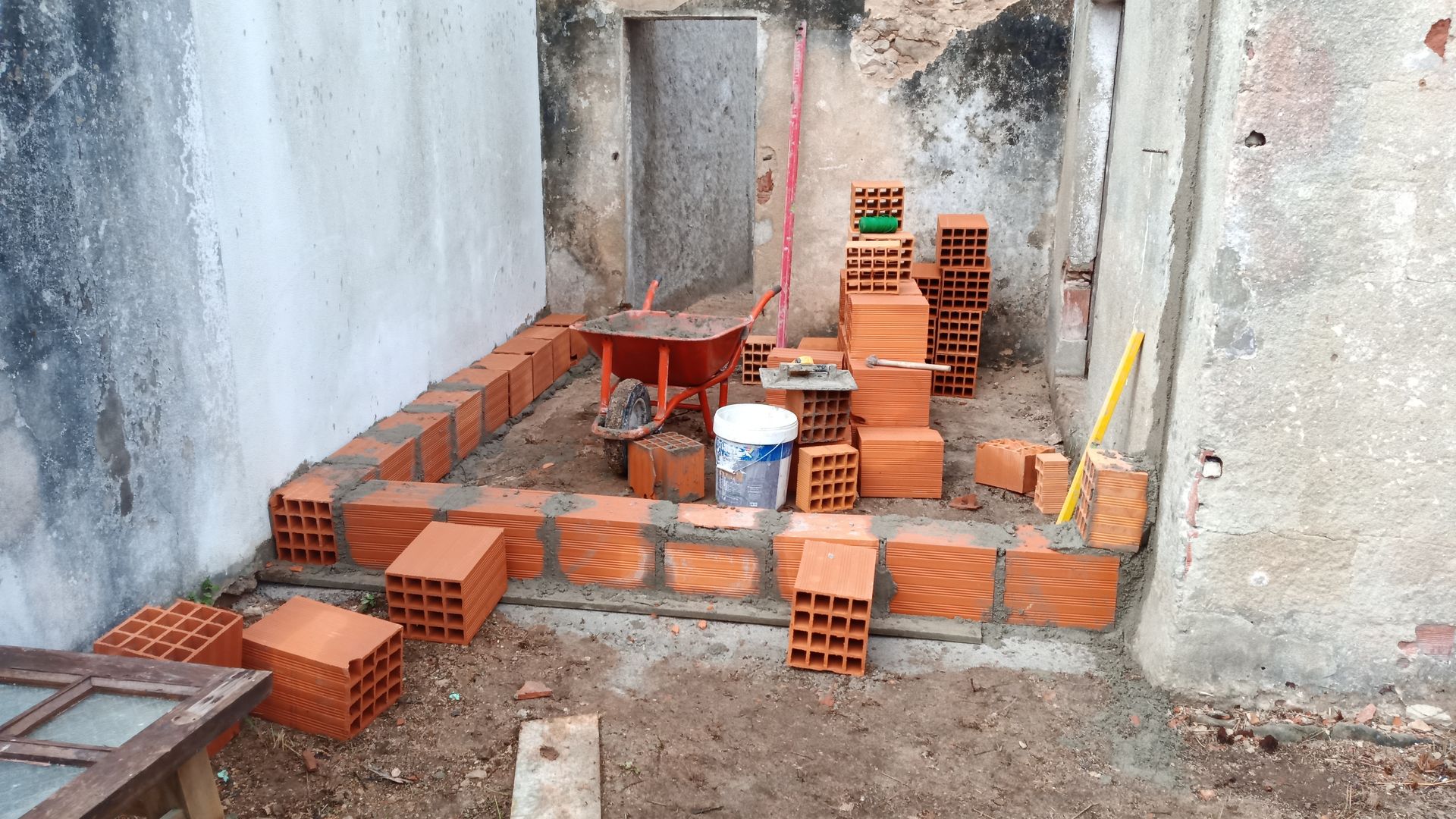 Canteiro de obras: Tijolos vermelhos sendo assentados para construir um pequeno muro em um espaço ao ar livre.