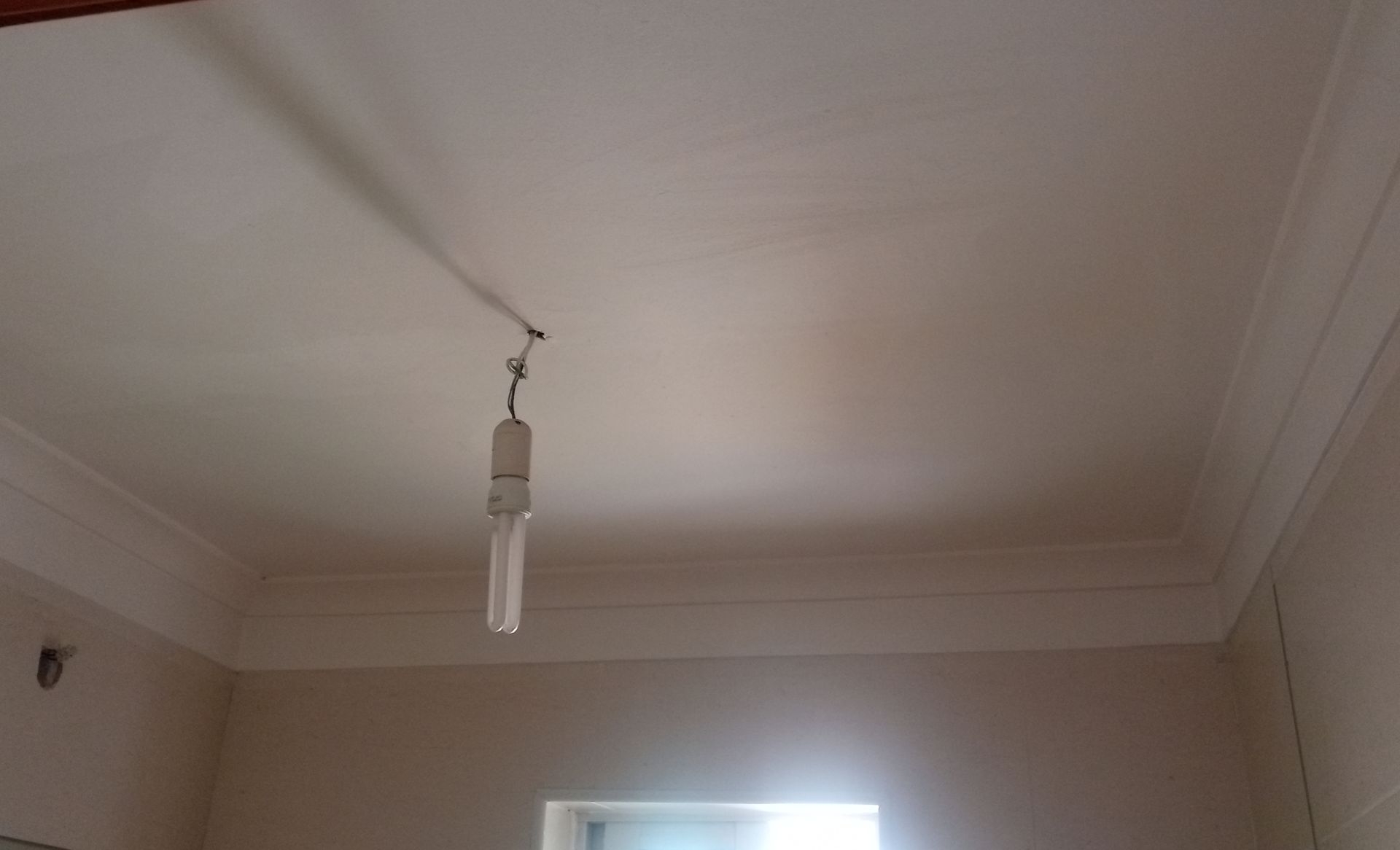 Teto branco com moldura decorativa. Uma lâmpada está pendurada por um fio no centro.