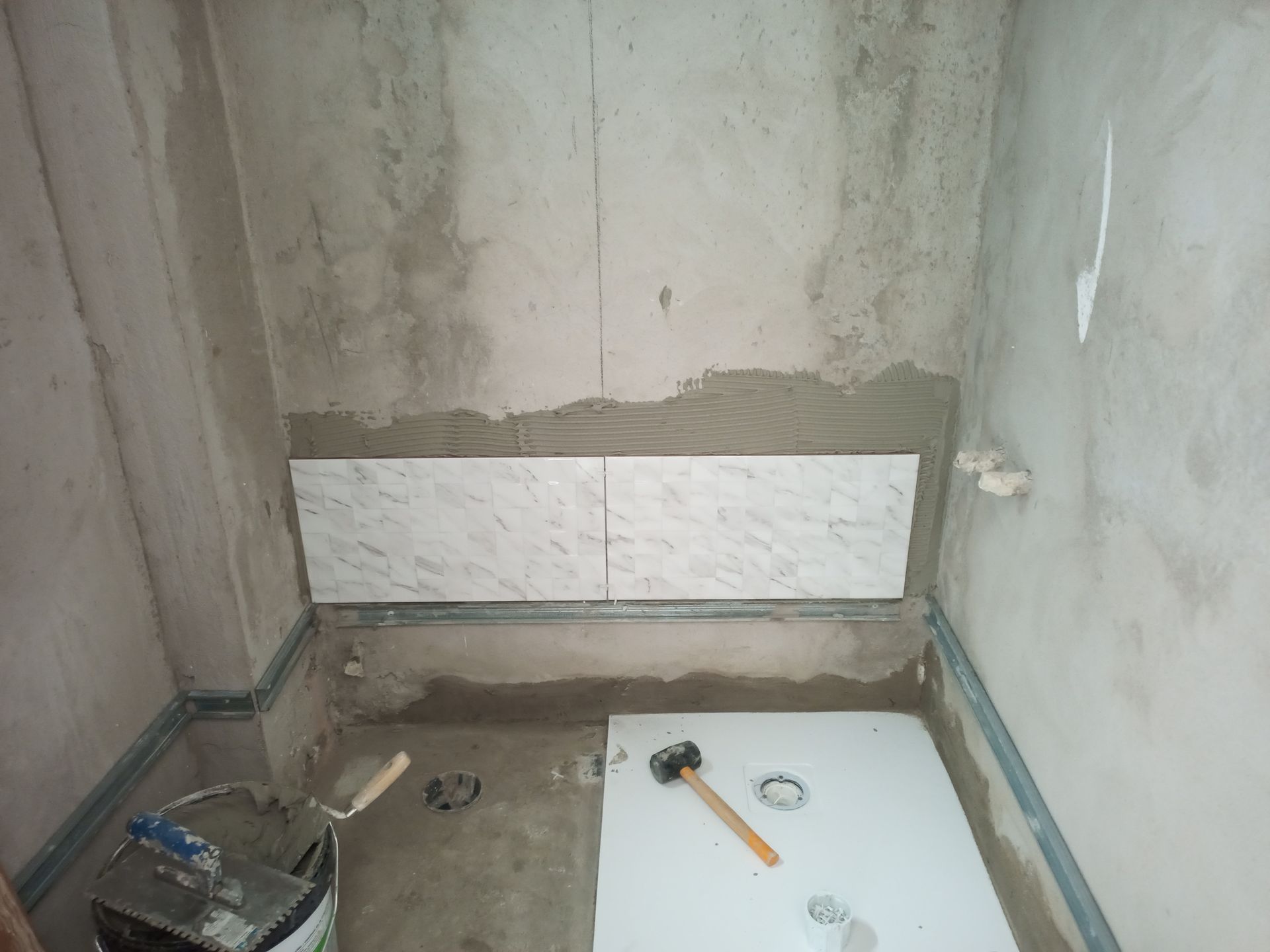 Banheiro em construção; azulejos parcialmente instalados na parede acima de uma prateleira, mostrando o acabamento em cimento e metal.