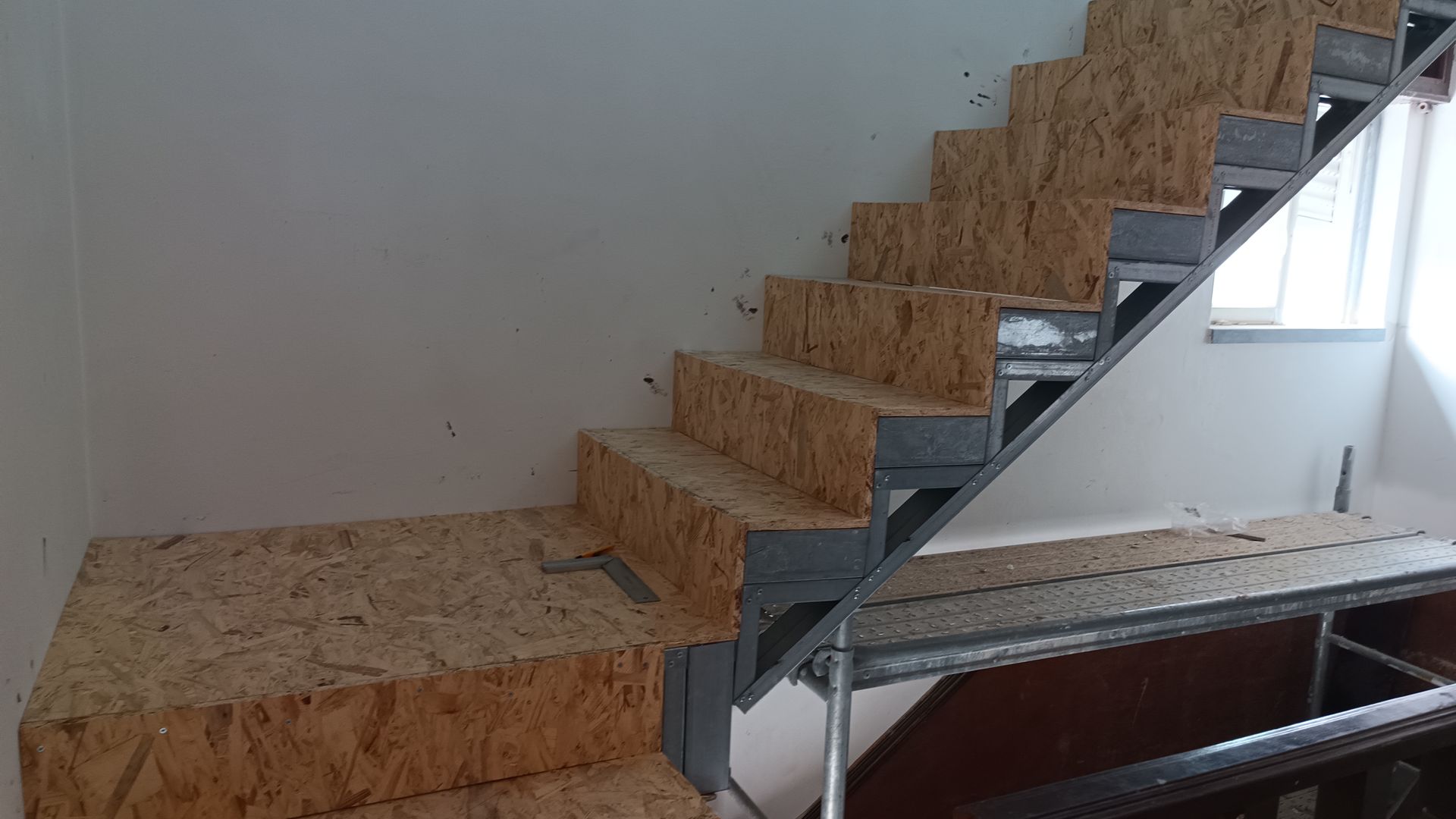 Escada interna inacabada, com estrutura de madeira e metal, encostada em uma parede branca.