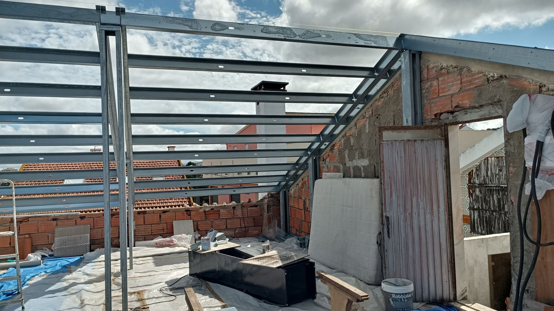 Estrutura metálica em construção no telhado, paredes de tijolo, céu nublado.